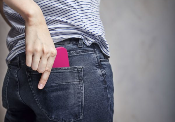 Main insérant un téléphone rose dans une poche de jean. L'arrière-plan est un mur uni.