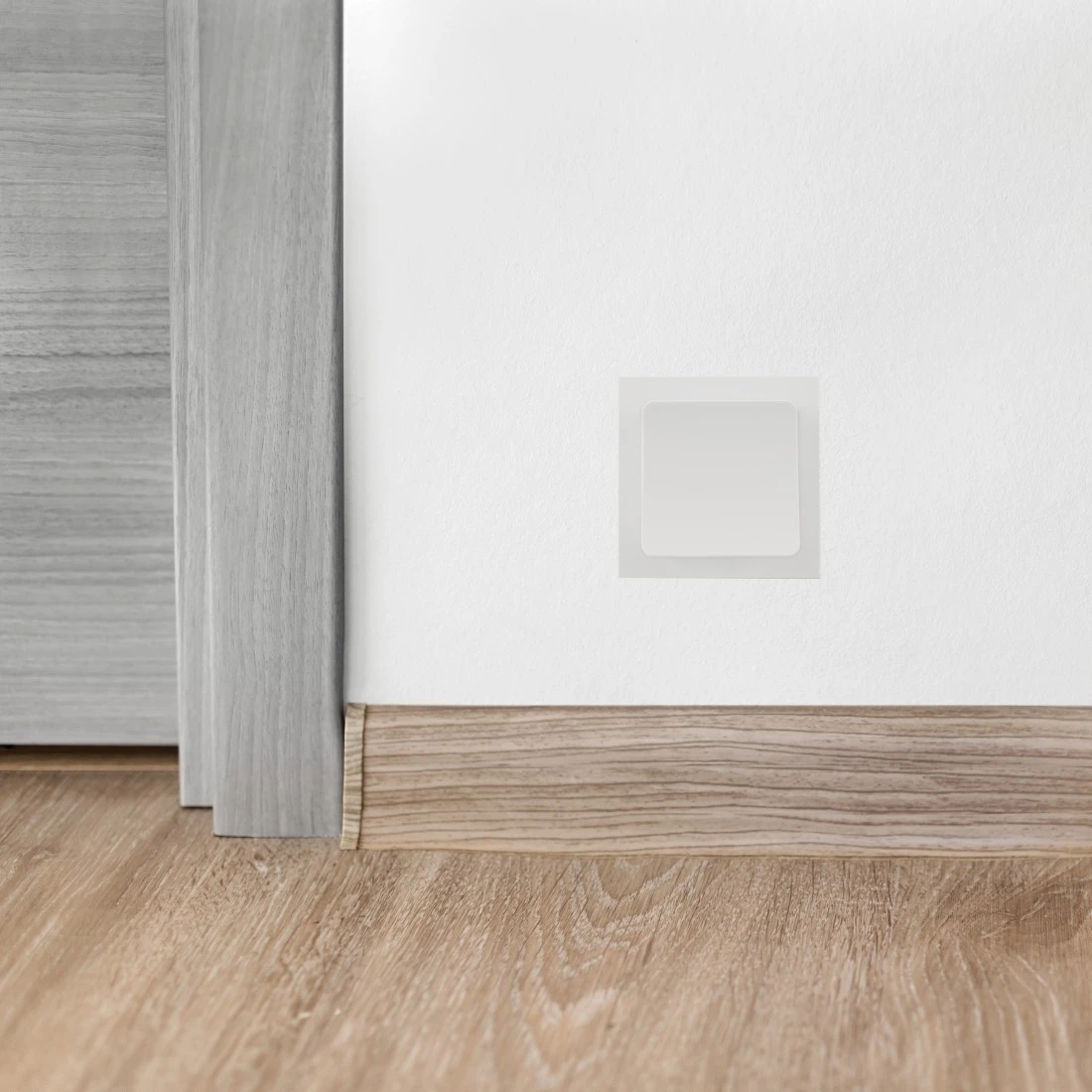 Muro bianco con copertura di ventilazione quadrata e di colore chiaro e pavimento in legno.