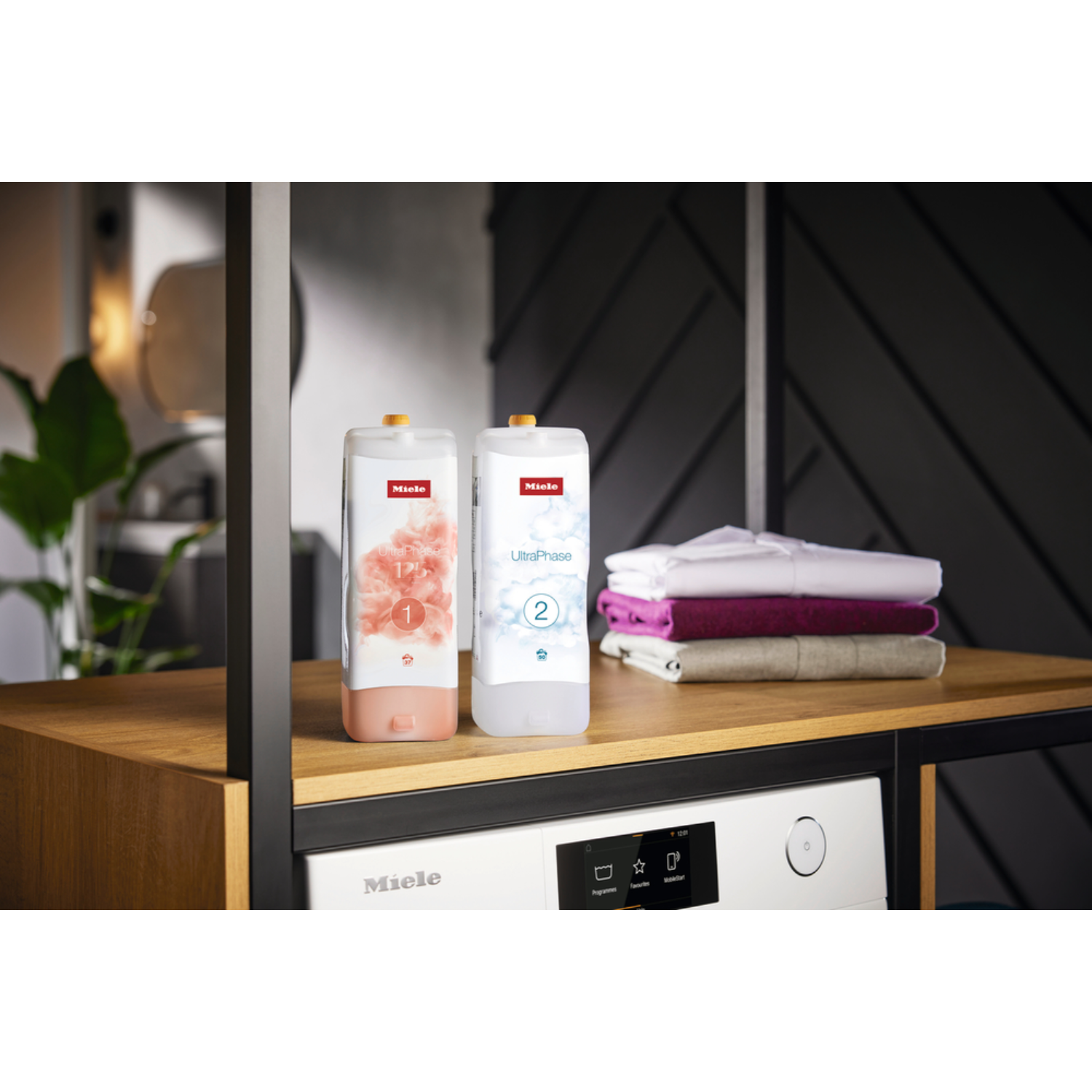 Two Miele UltraPhase laundry detergent bottles and folded towels on a wooden shelf. A washing machine is below.