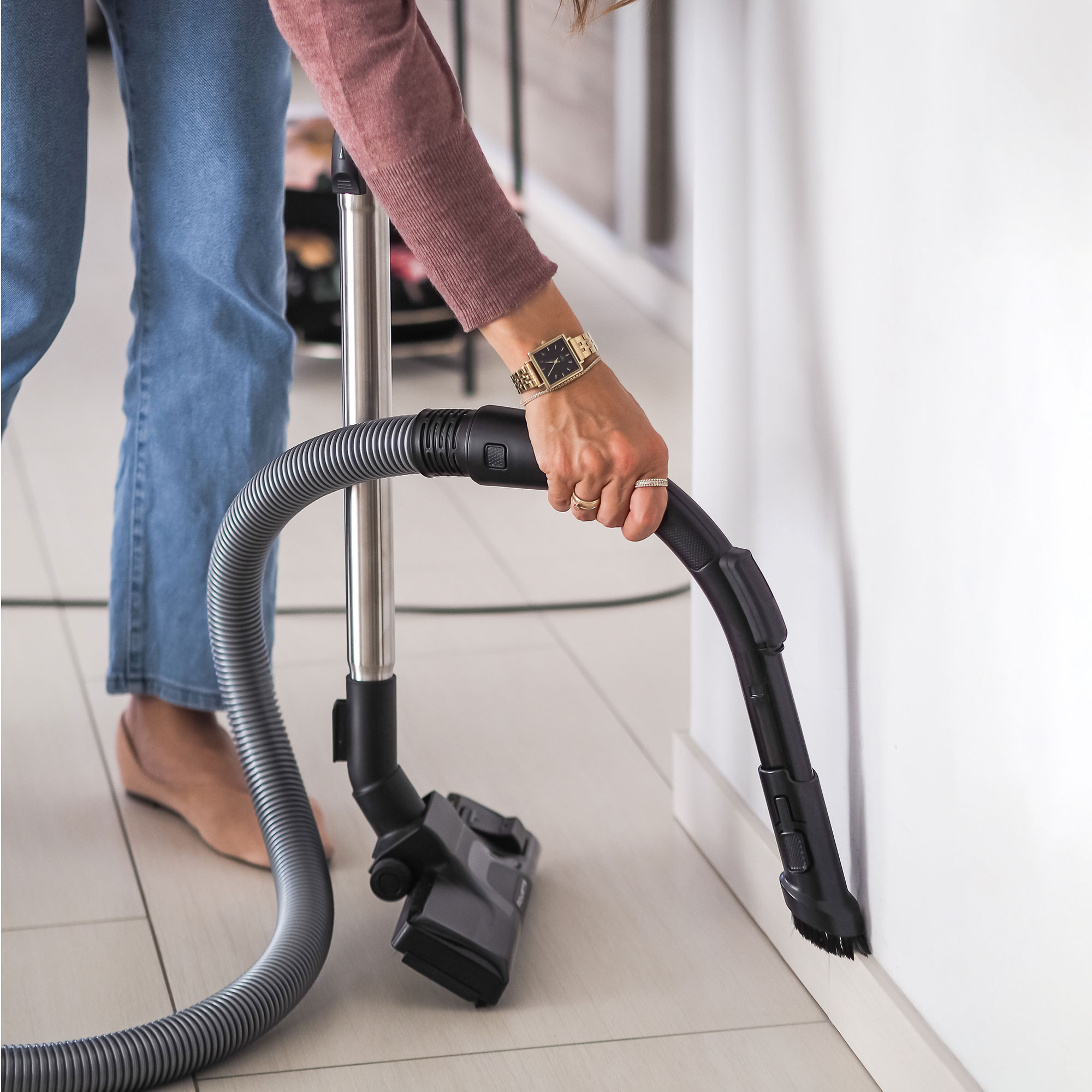 Mujer en jeans aspira una esquina. Lleva reloj y anillos. La aspiradora es negra y gris.