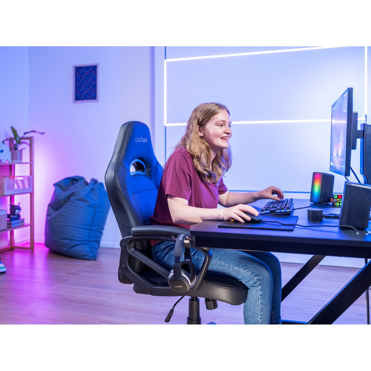 Une jeune femme souriante joue sur un bureau. Un écran et des haut-parleurs sont visibles.
