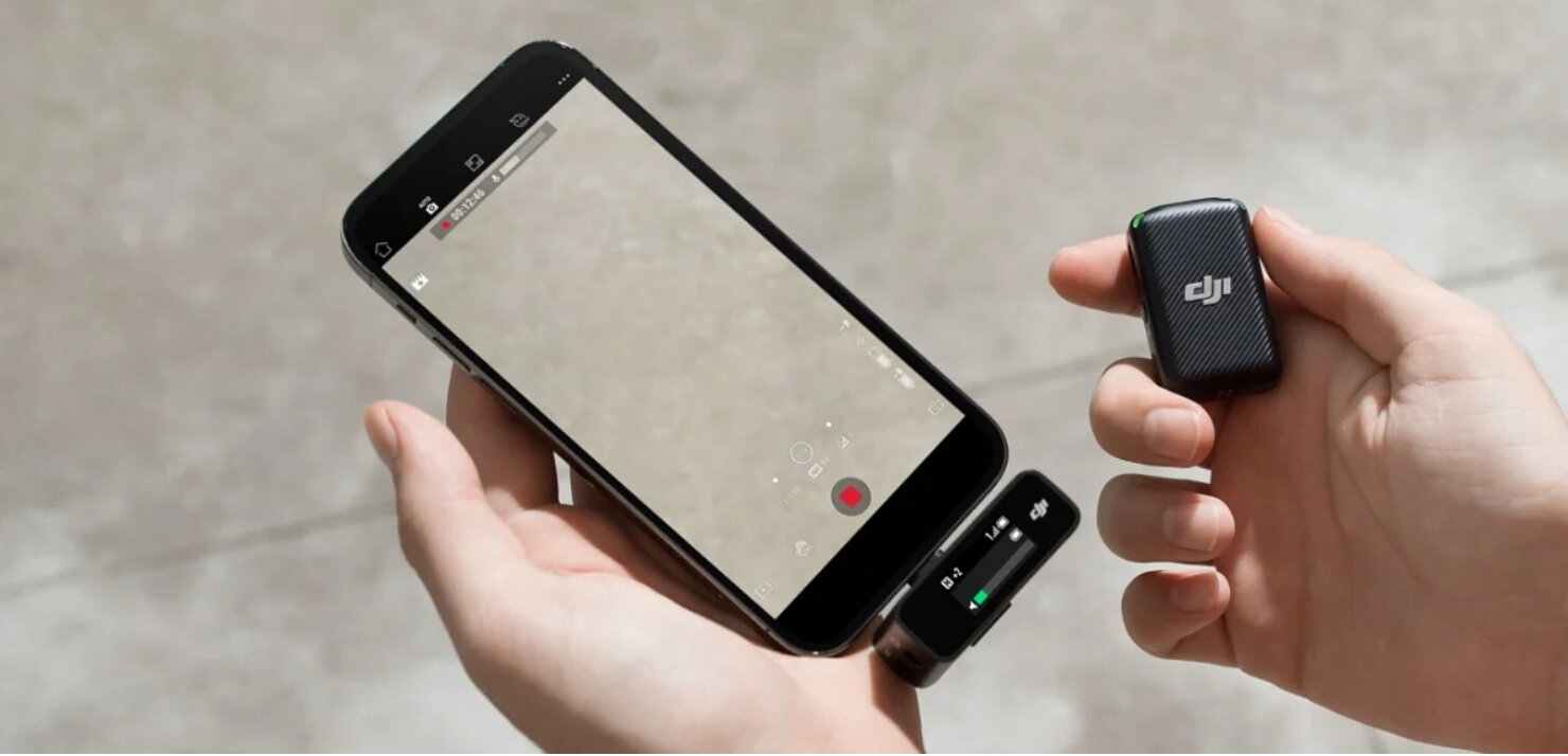 Osoba trzymająca telefon i dwa czarne mikrofony bezprzewodowe, na rozmytym tle.