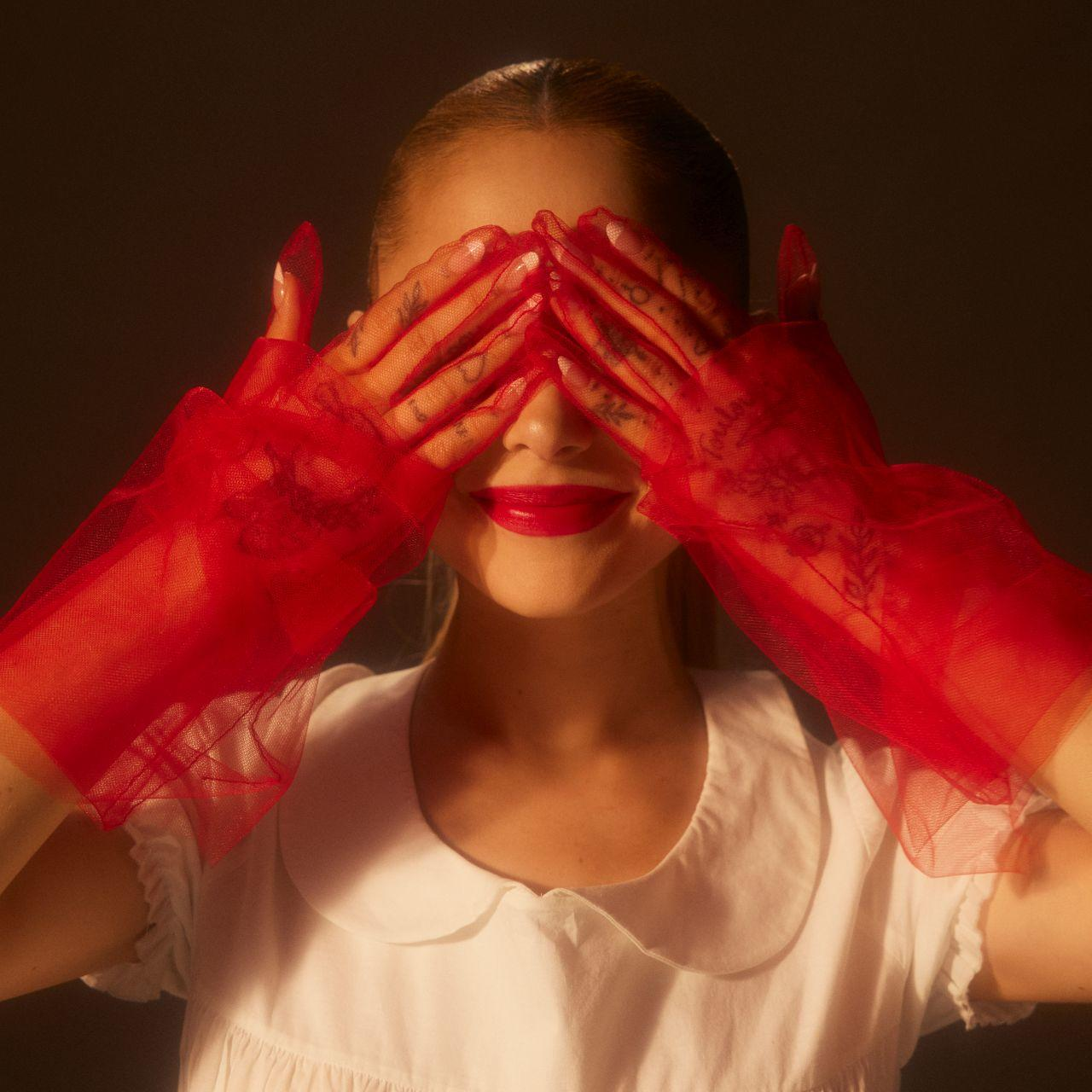 Frau mit roten Handschuhen, die ihre Augen bedecken, trägt ein weißes Kleid, dunkler Hintergrund.
