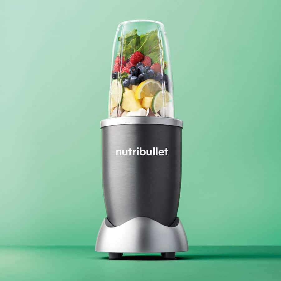 Egy Nutribullet turmixgép gyümölcsökkel és zöldségekkel, zöld háttér előtt.