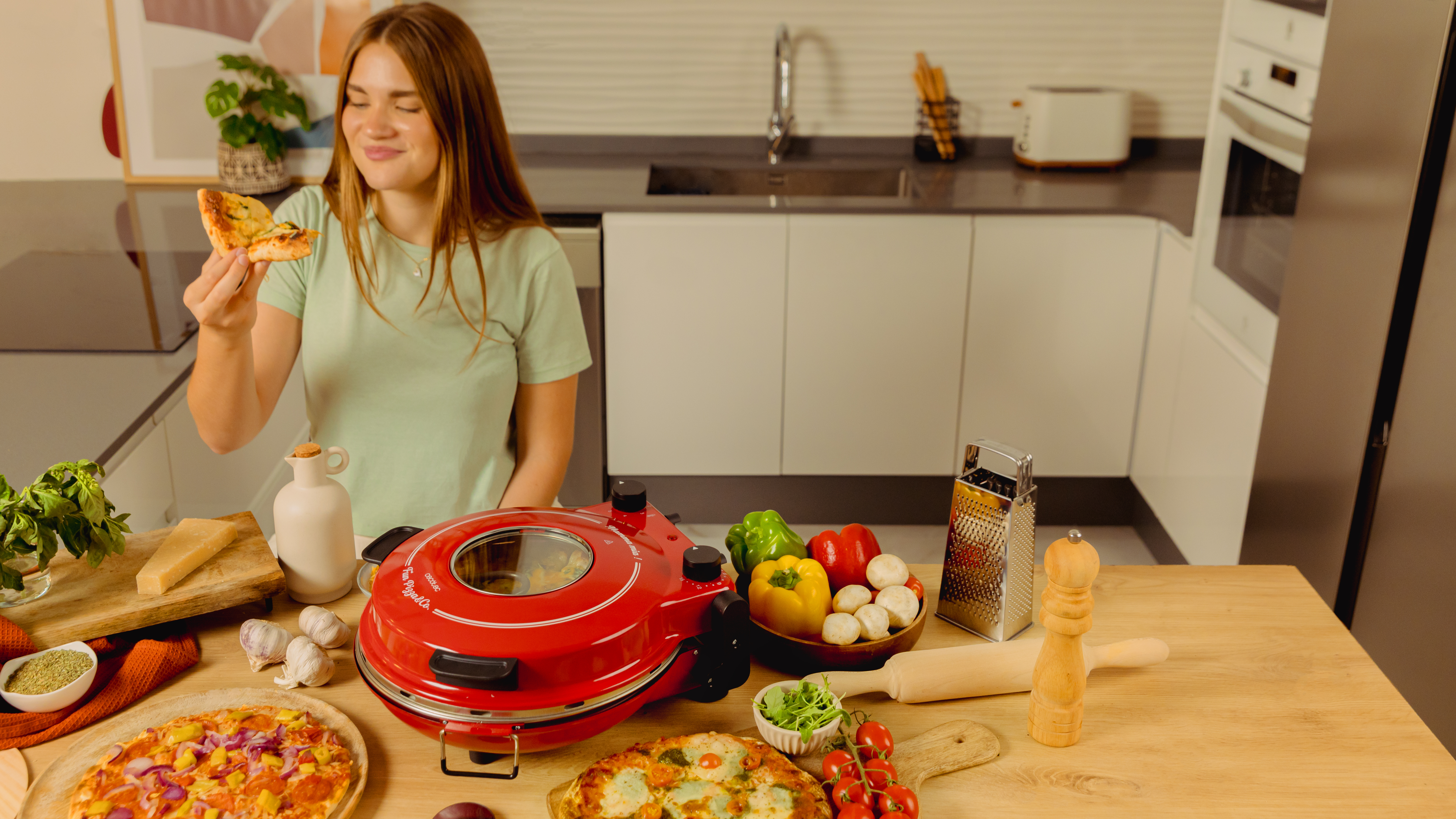 Mujer comiendo porción de pizza, ambiente de cocina. Horno, ingredientes y comida en mesa.