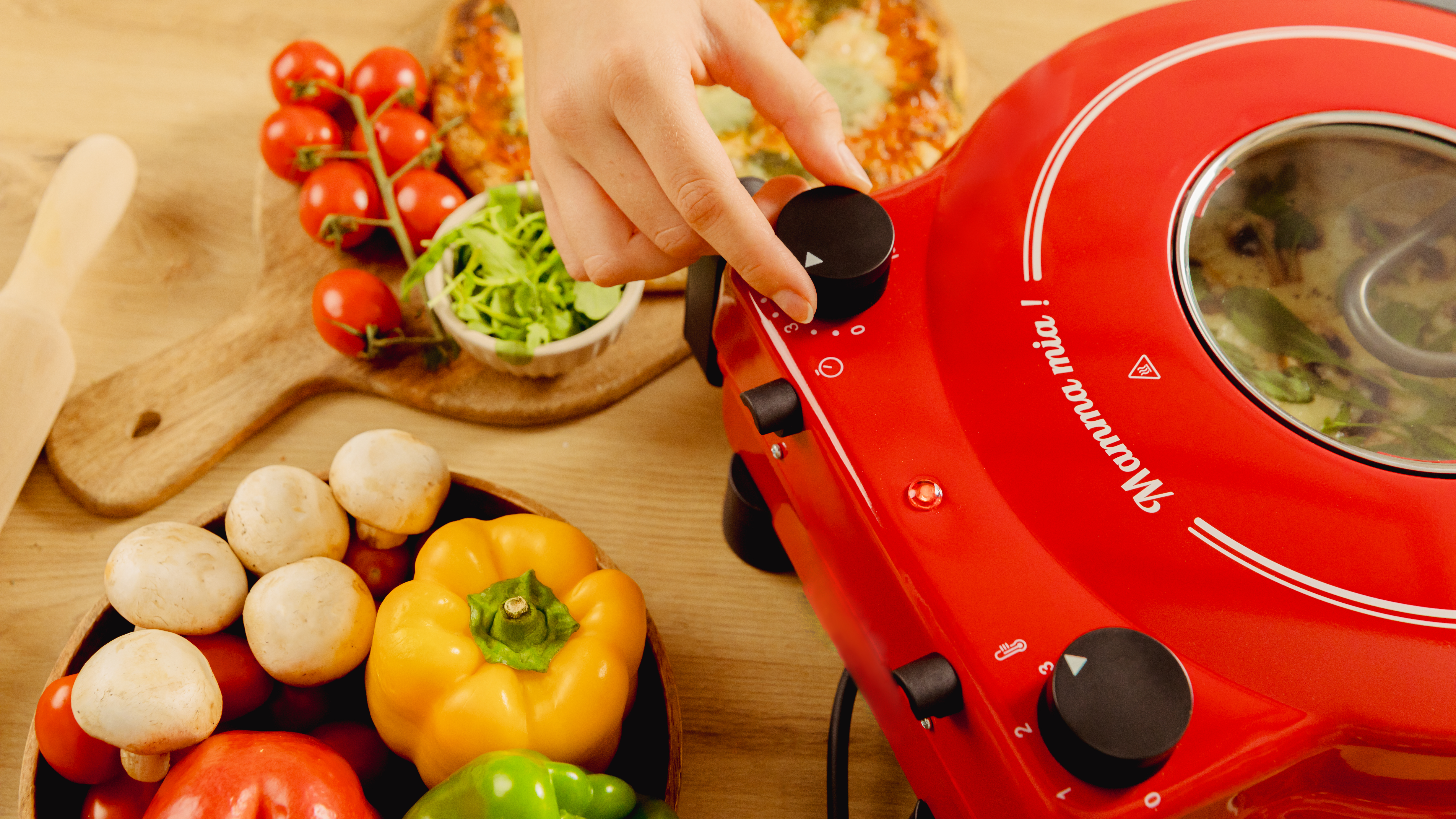 Una mano gira una perilla en un horno de pizza rojo. Verduras y pizza están en la mesa de madera.