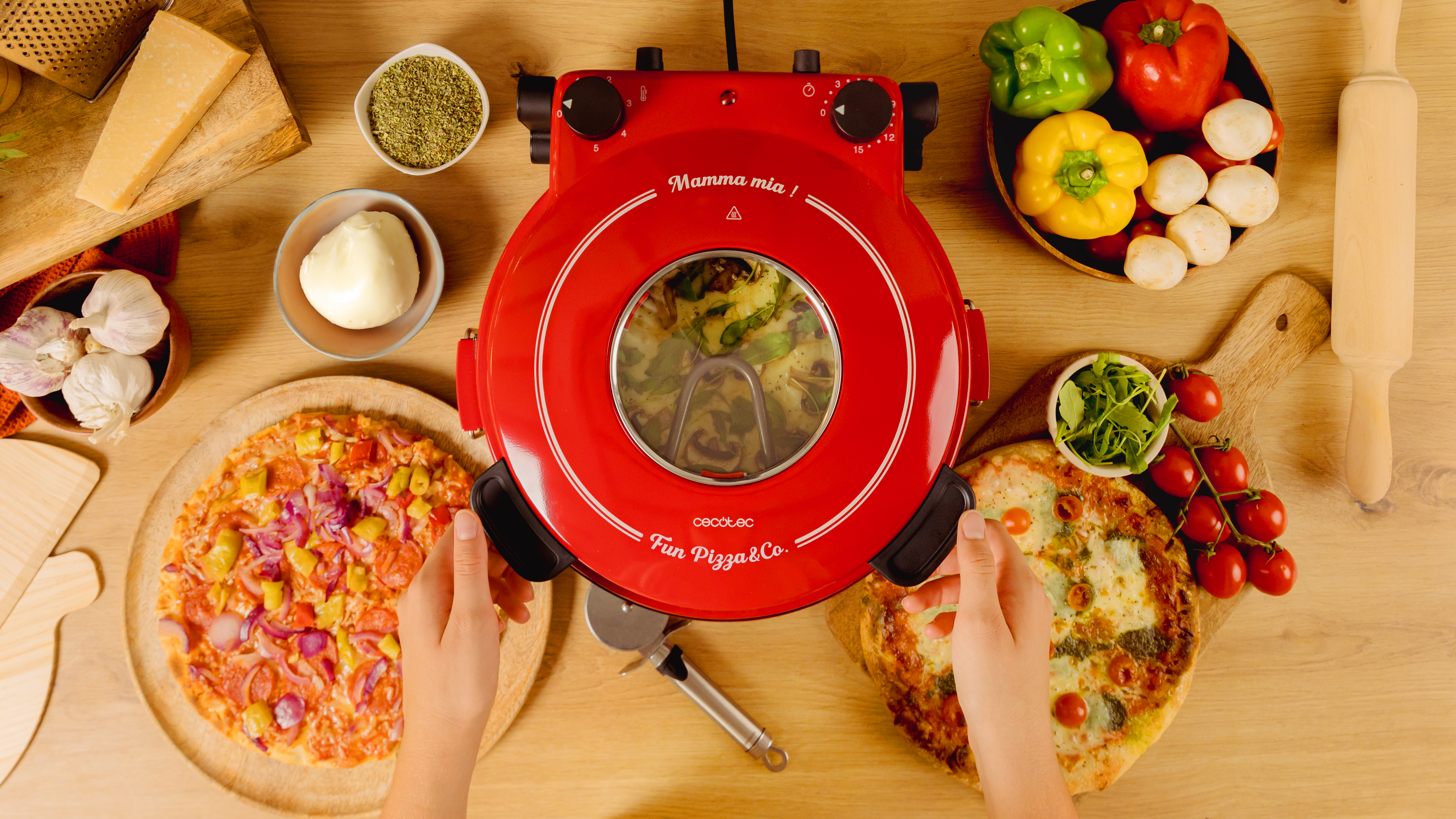 Un horno de pizza rojo con pizzas e ingredientes sobre una mesa de madera. Dos manos sostienen pizza.