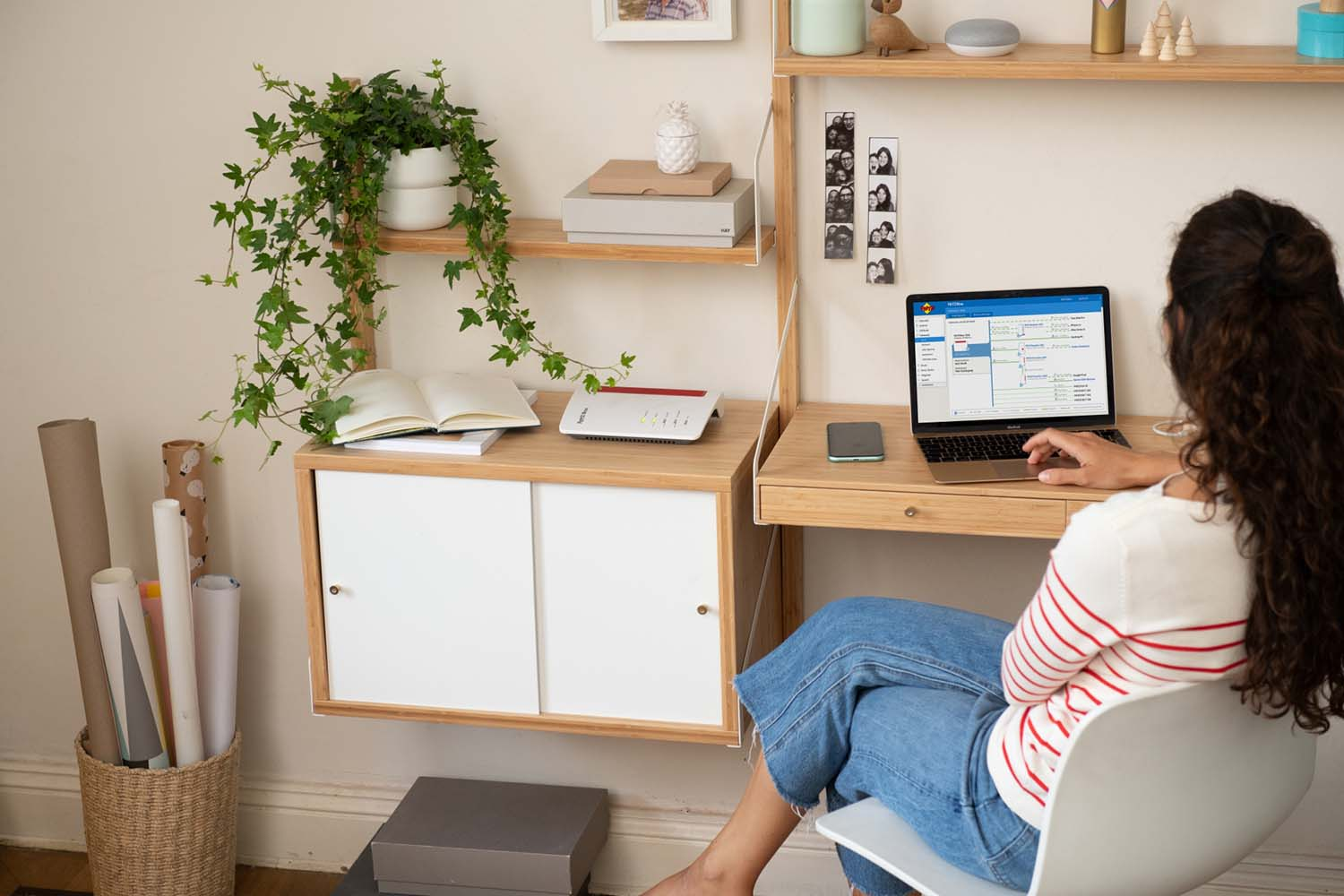 Mujer trabajando en un portátil en un escritorio con una planta, libros y un enrutador. Estanterías de pared.
