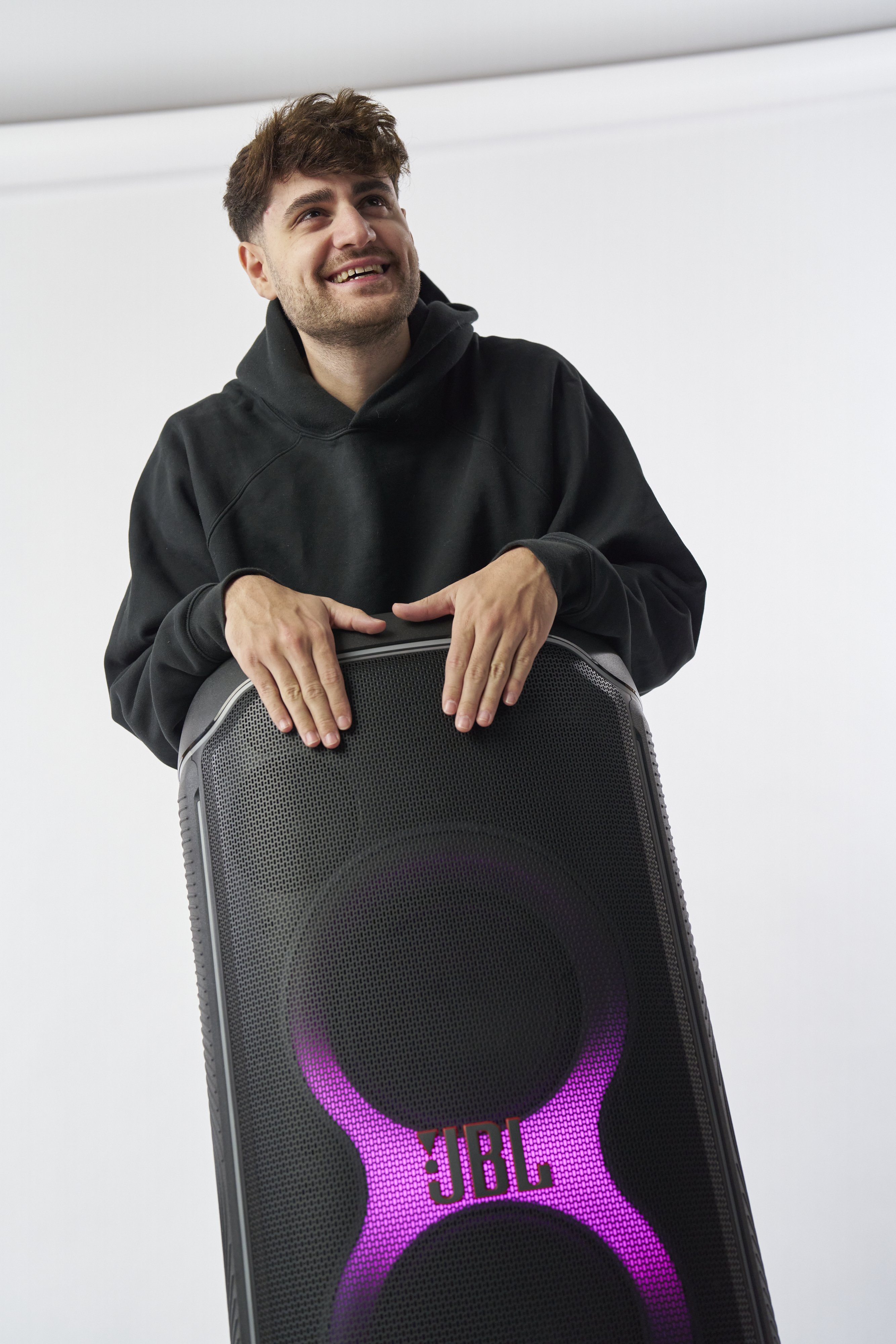 Mann mit schwarzem Kapuzenpullover lächelt neben einem JBL-Lautsprecher mit lila Lichtern.