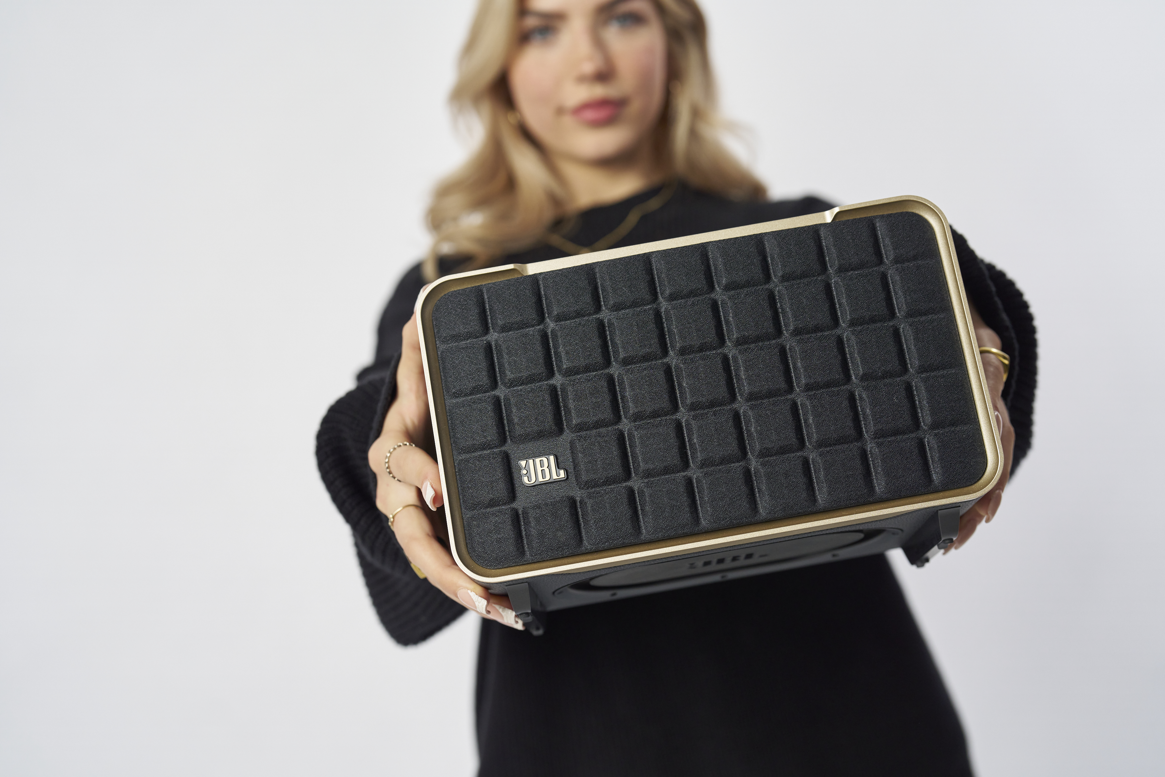 Une femme tient un haut-parleur noir et or, le logo JBL est visible.