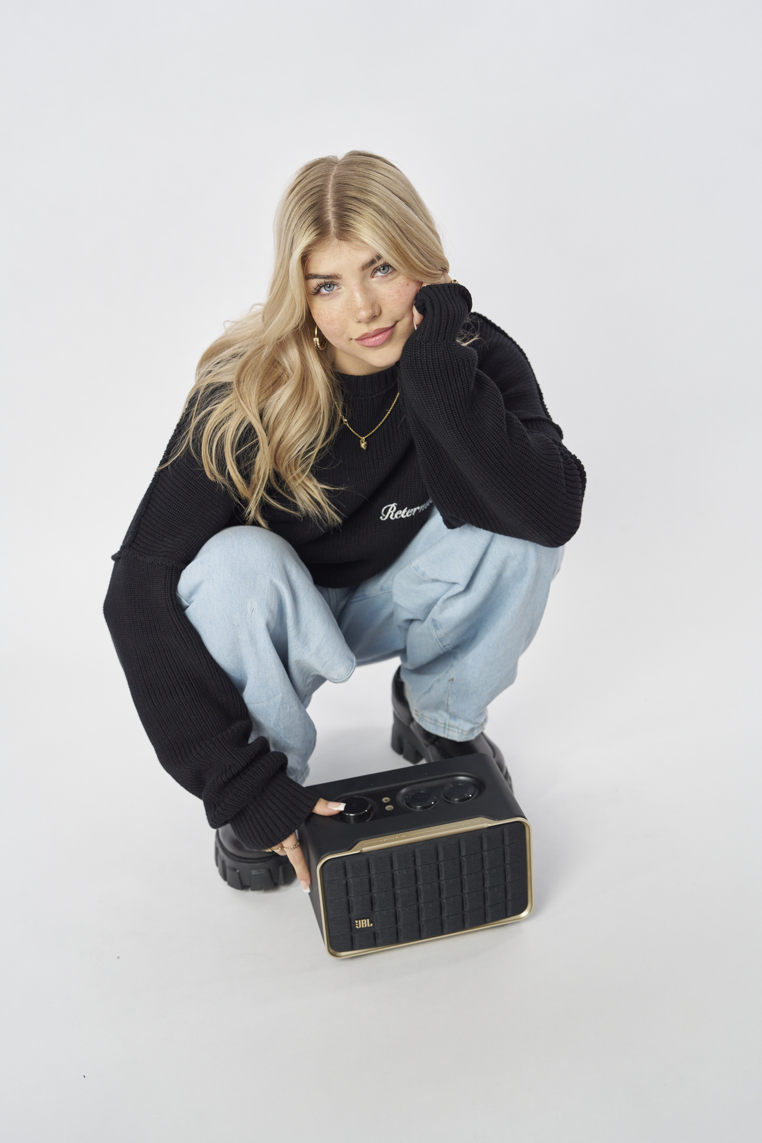 Une femme accroupie avec un haut-parleur JBL noir, vêtue d'un pull noir et d'un jean.
