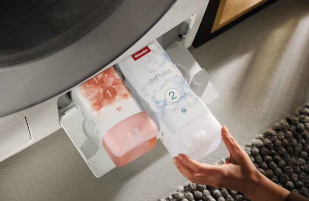 Person fügt Waschmittel in Waschmaschine ein. Zwei Waschmittelpackungen im Spender.
