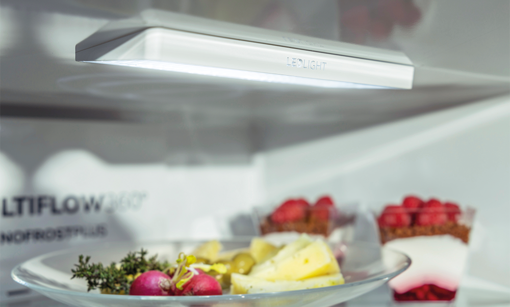 Wewnątrz lodówki, jasne światło LED oświetla półki z jedzeniem, w tym talerz z jedzeniem i deserami.