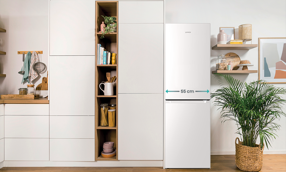 Biała lodówka, szeroka na 55 cm, w kuchni z drewnianymi półkami i szafkami. Roślina po prawej.