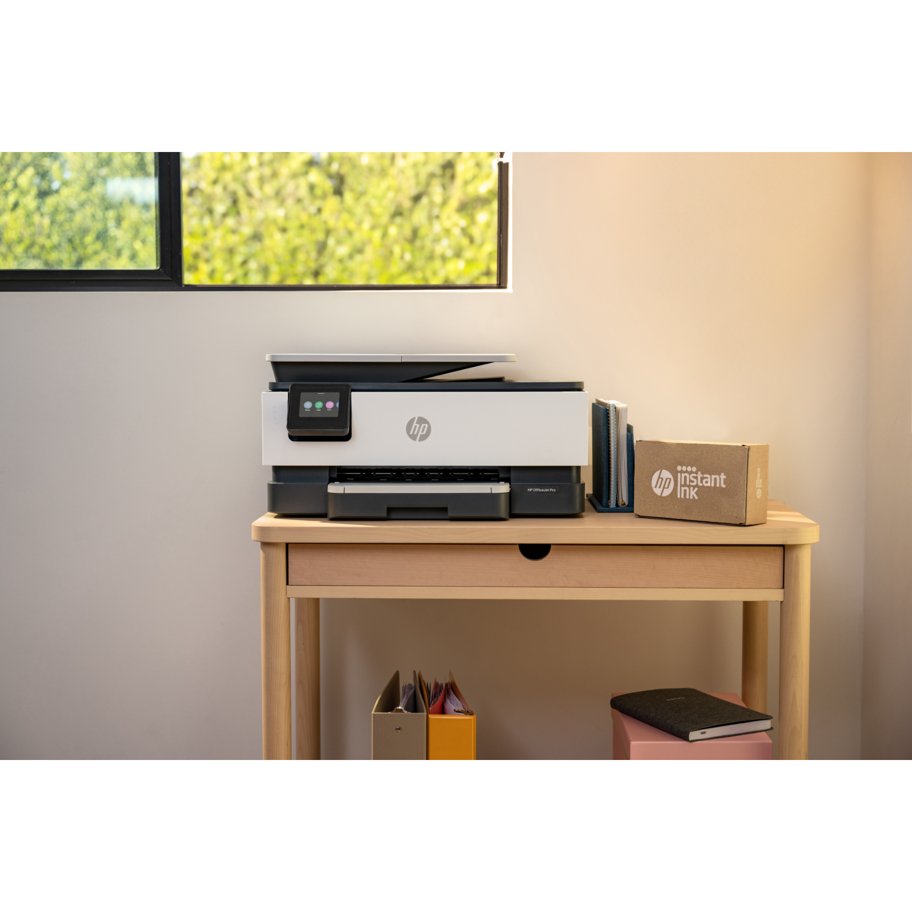 White printer, desk with books, notepad, and instant ink box. A window shows a green bush outside.