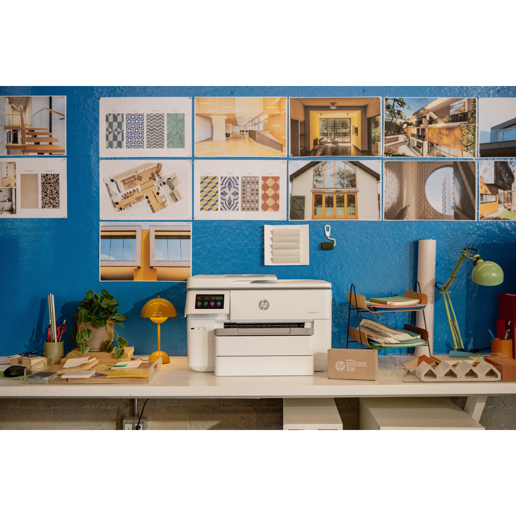 Een bureau met een printer en meerdere items, waaronder een plant, lamp en kunstwerk, tegen een blauwe muur.