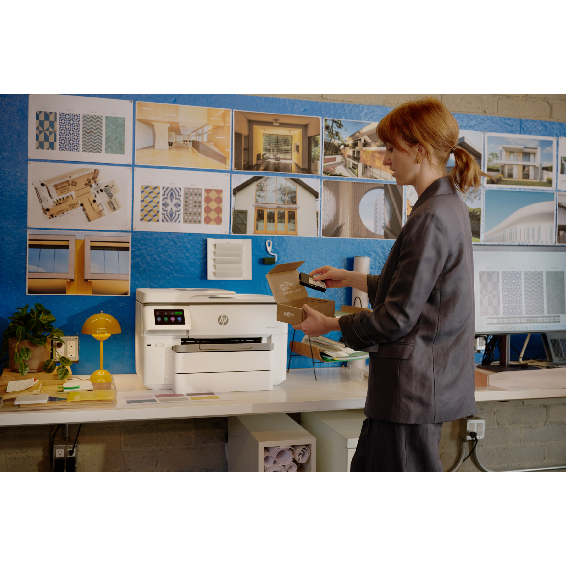 Vrouw houdt een inktbox vast bij een witte printer op een bureau. Architectuurontwerpen op blauwe muur.