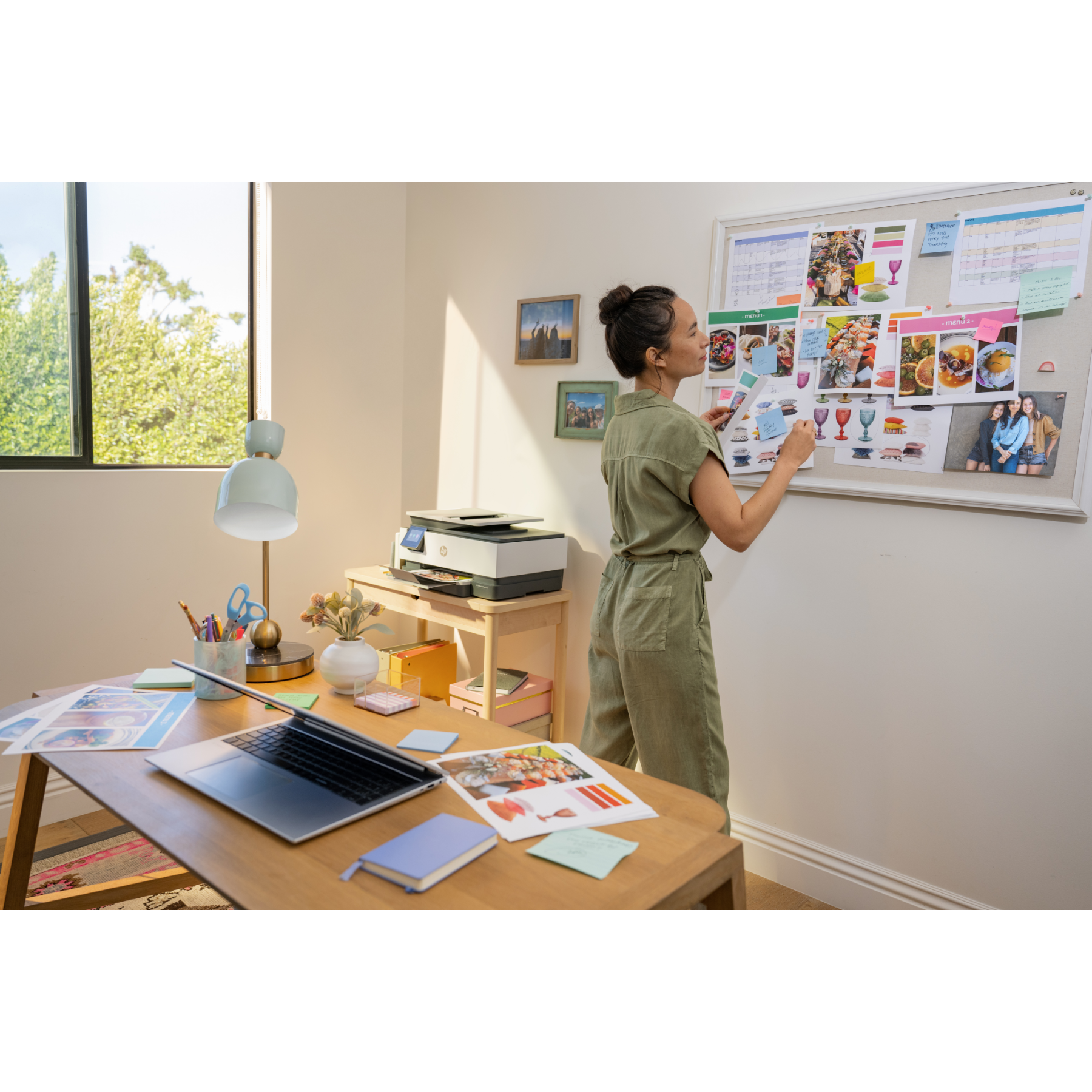 Vrouw bij een whiteboard met papieren, bureau met laptop en printer, raam op de achtergrond.