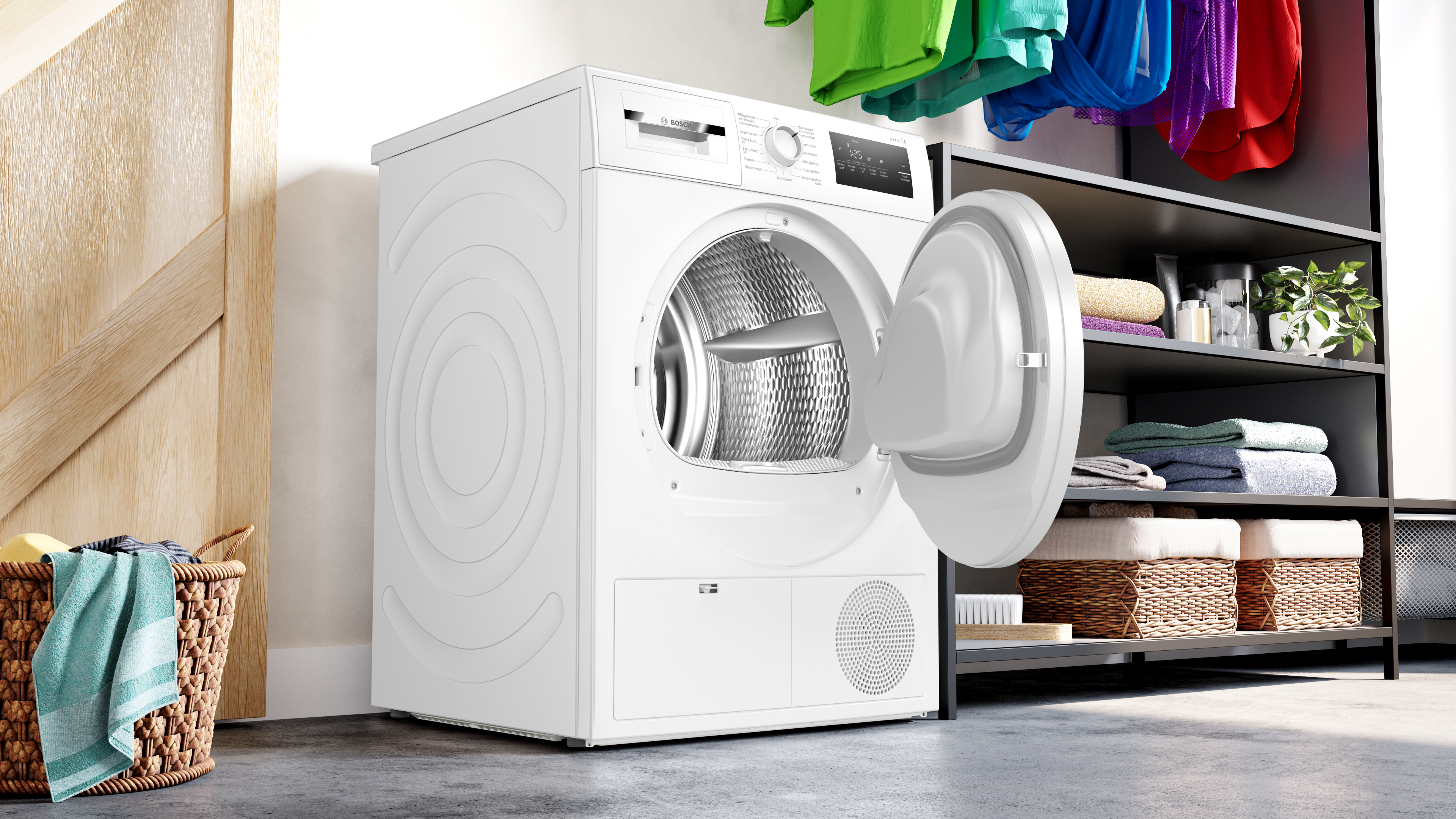Eine weiße Waschmaschine mit offener Tür. Bunte Kleidung hängt darüber. Ein Wäschekorb steht auf dem Boden.