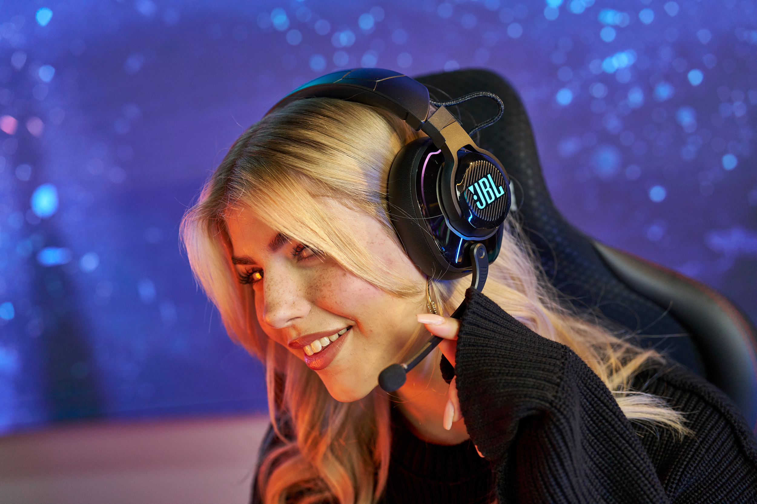 Une femme souriante portant un casque JBL avec micro, jouant dans une pièce bleue.
