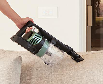Hand hält einen schwarz-grünen Staubsauger, der ein beiges Sofa reinigt.