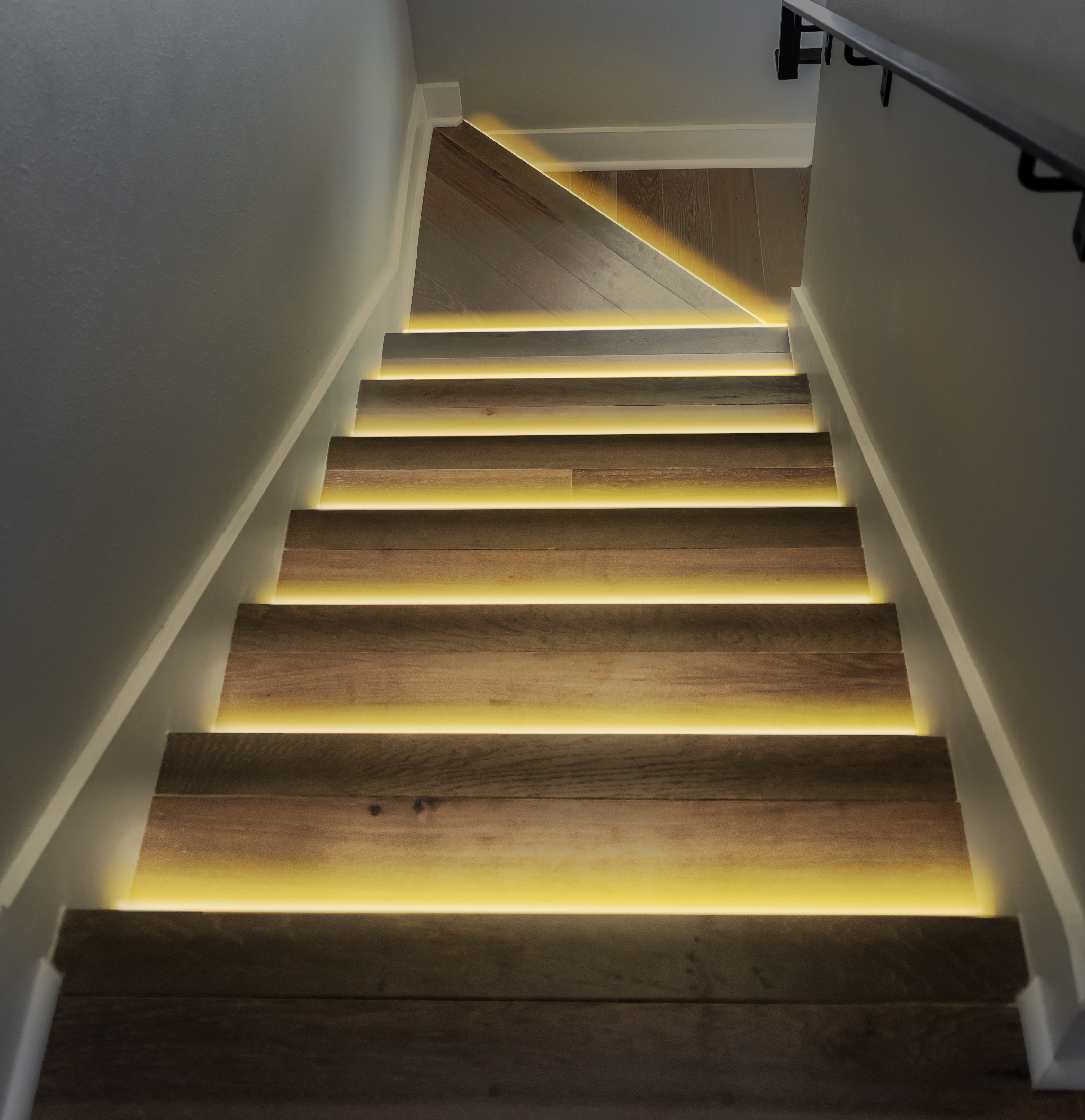 Holztreppe mit gelben Lichtern. Weiße Wände und ein schwarzer Handlauf sind sichtbar.