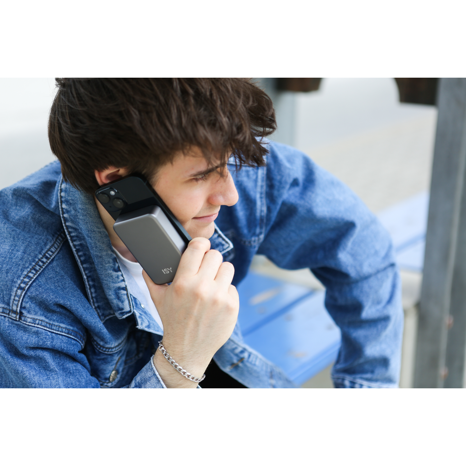 Een jonge man houdt een telefoon tegen zijn oor terwijl hij een spijkerjack en een wit shirt draagt. Hij is buiten.