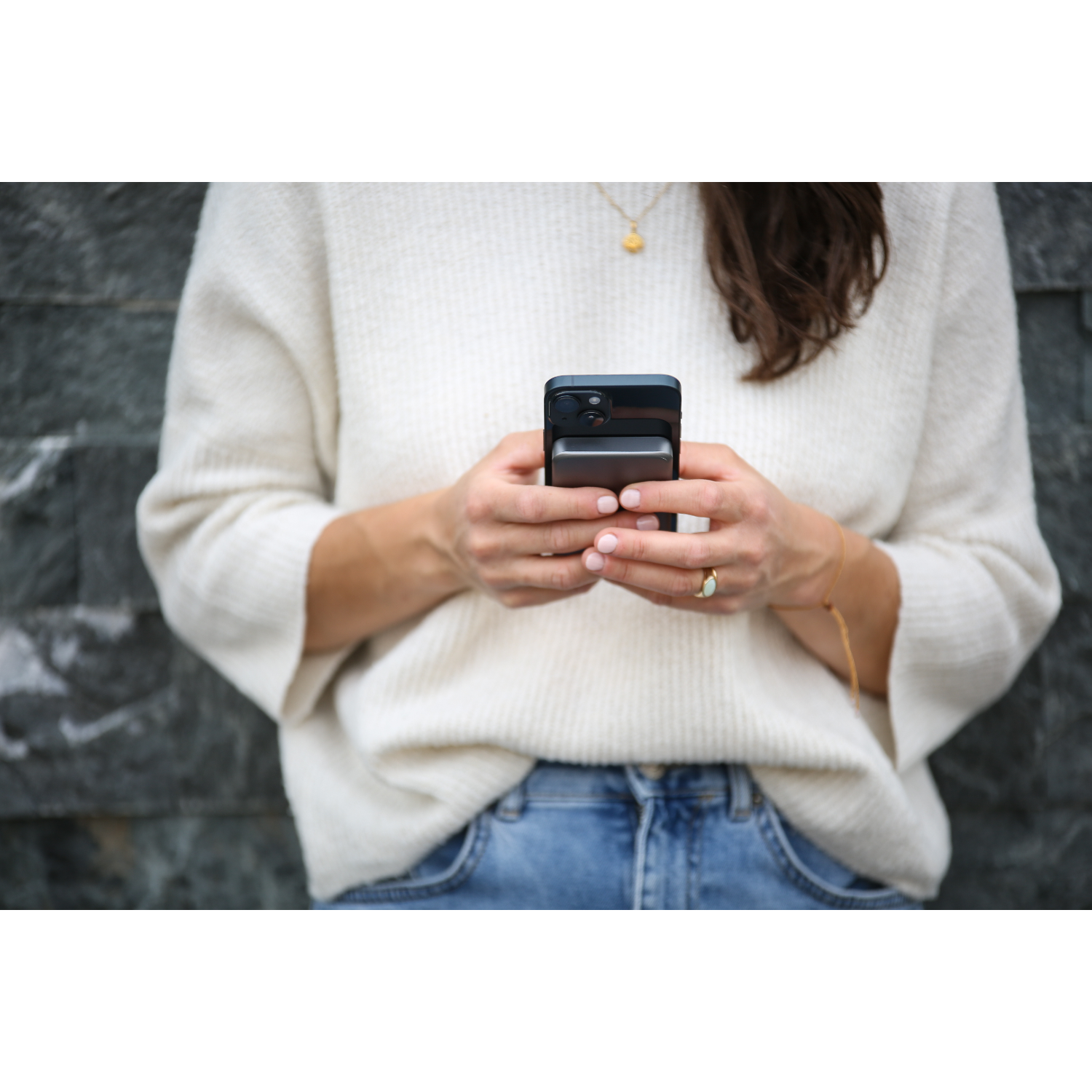 A person holding a phone and a power bank. The person wears a white sweater and jeans. The background is a stone wall.