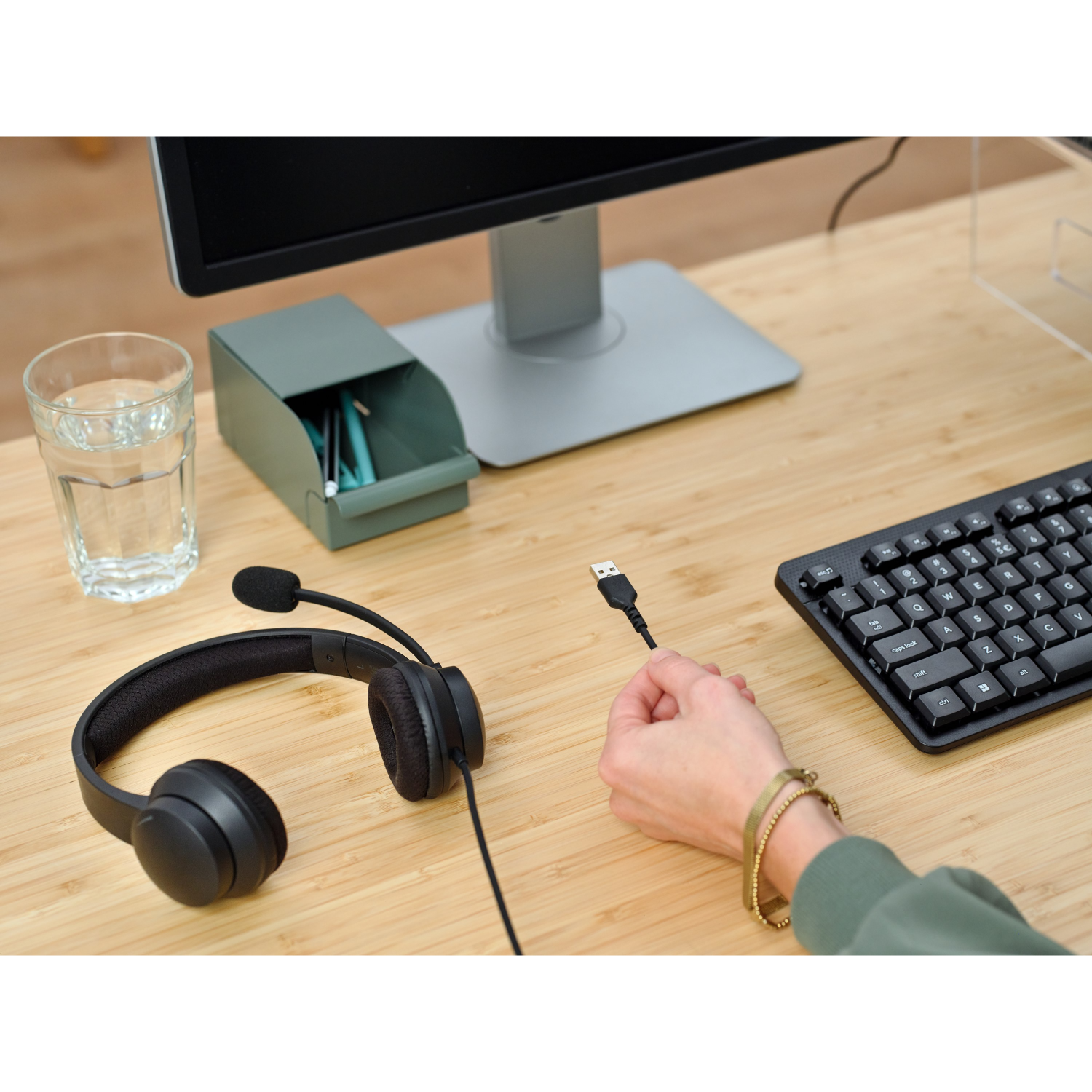 A desk with a headset and keyboard. A person's hand holds a USB cable. A glass of water is also on the desk.
