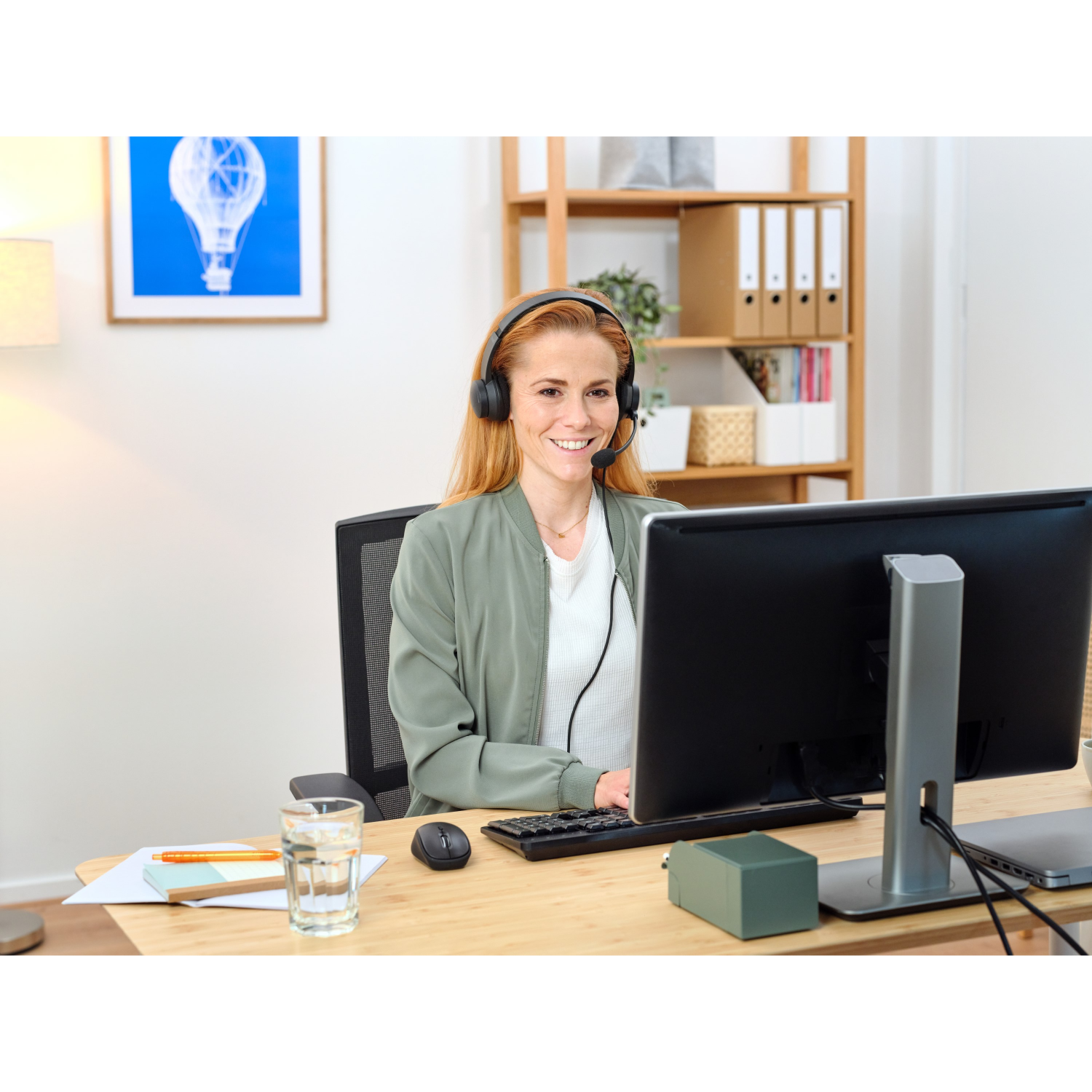 A woman with a headset sits at a desk, working on a computer.