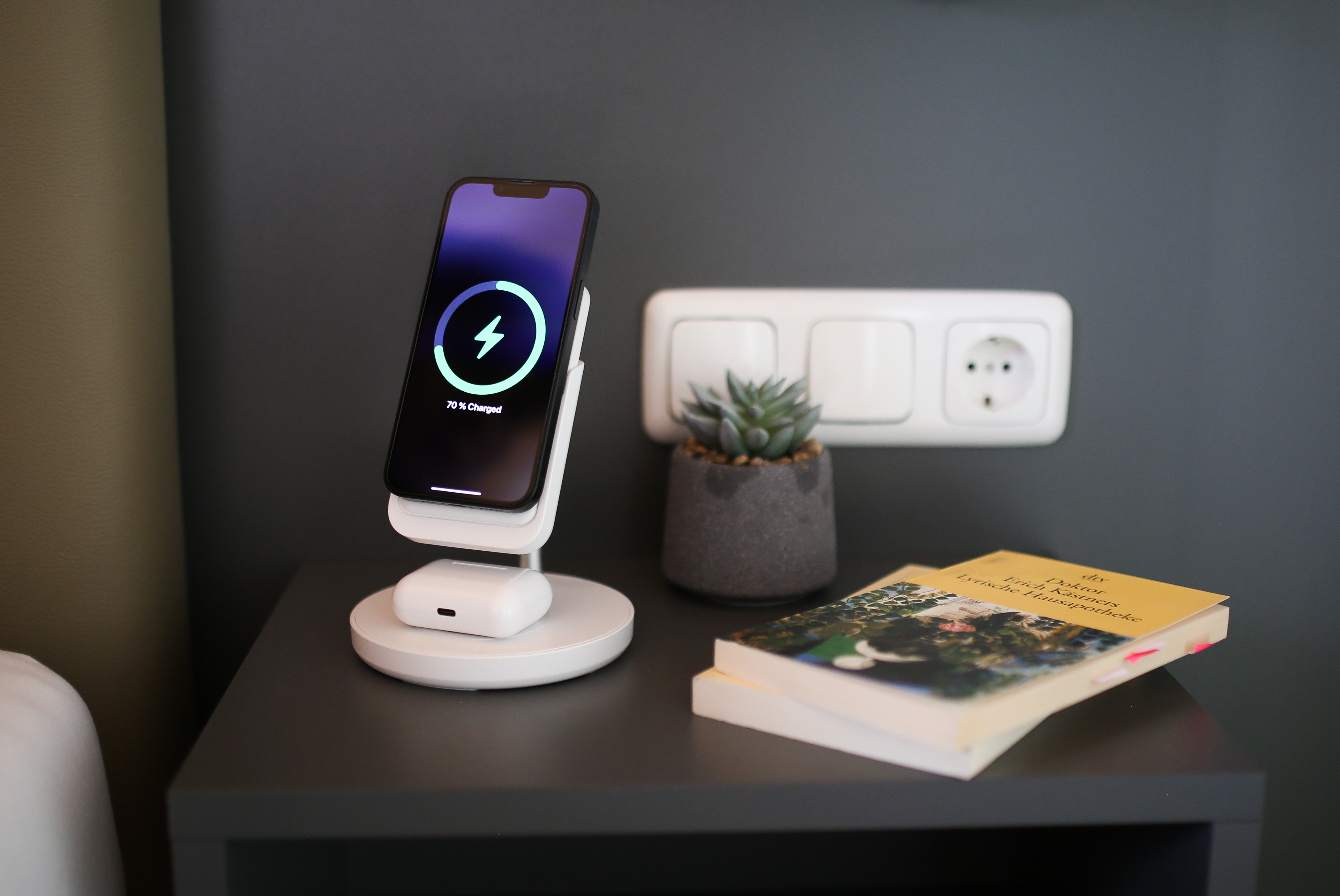 Ein Telefon, das drahtlos auf einem weißen Ladeständer geladen wird. Eine Pflanze und ein Buch liegen auf dem Tisch.