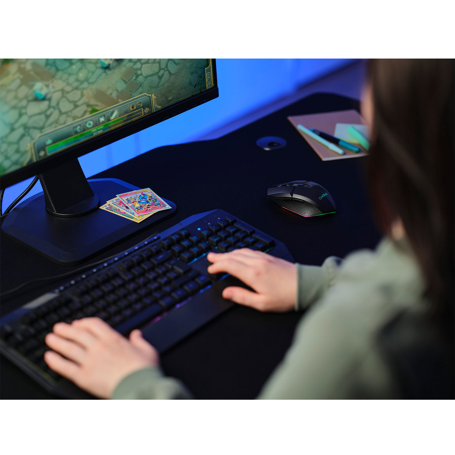 Persona usa la tastiera. Monitor mostra il gioco. Mouse a destra. Scrivania nera. Carte sulla scrivania.