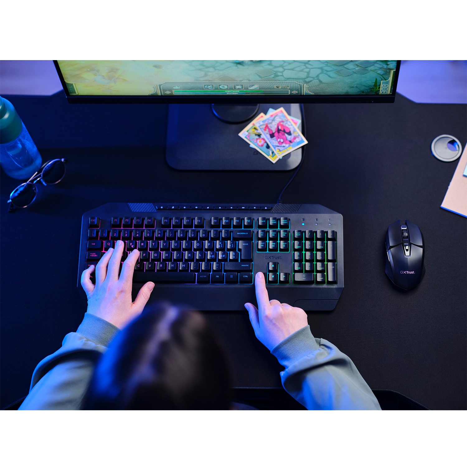 Persona che utilizza una tastiera retroilluminata, con un mouse, monitor e altri oggetti su una scrivania scura.