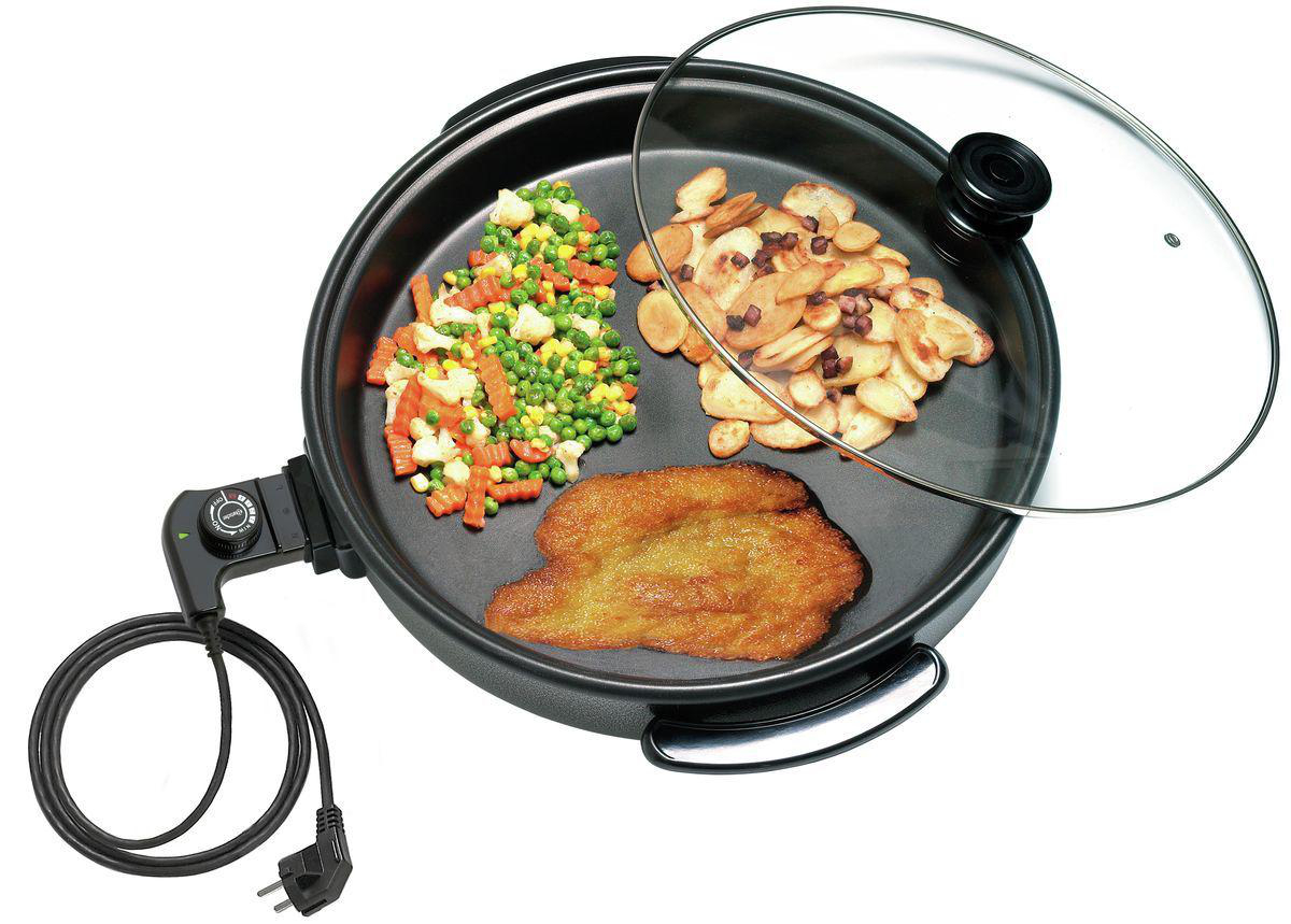 Elektrische Bratpfanne mit Gemüse, Kartoffeln und einem Stück gebratenem Fleisch. Ein Glasdeckel bedeckt einen Teil der Pfanne.