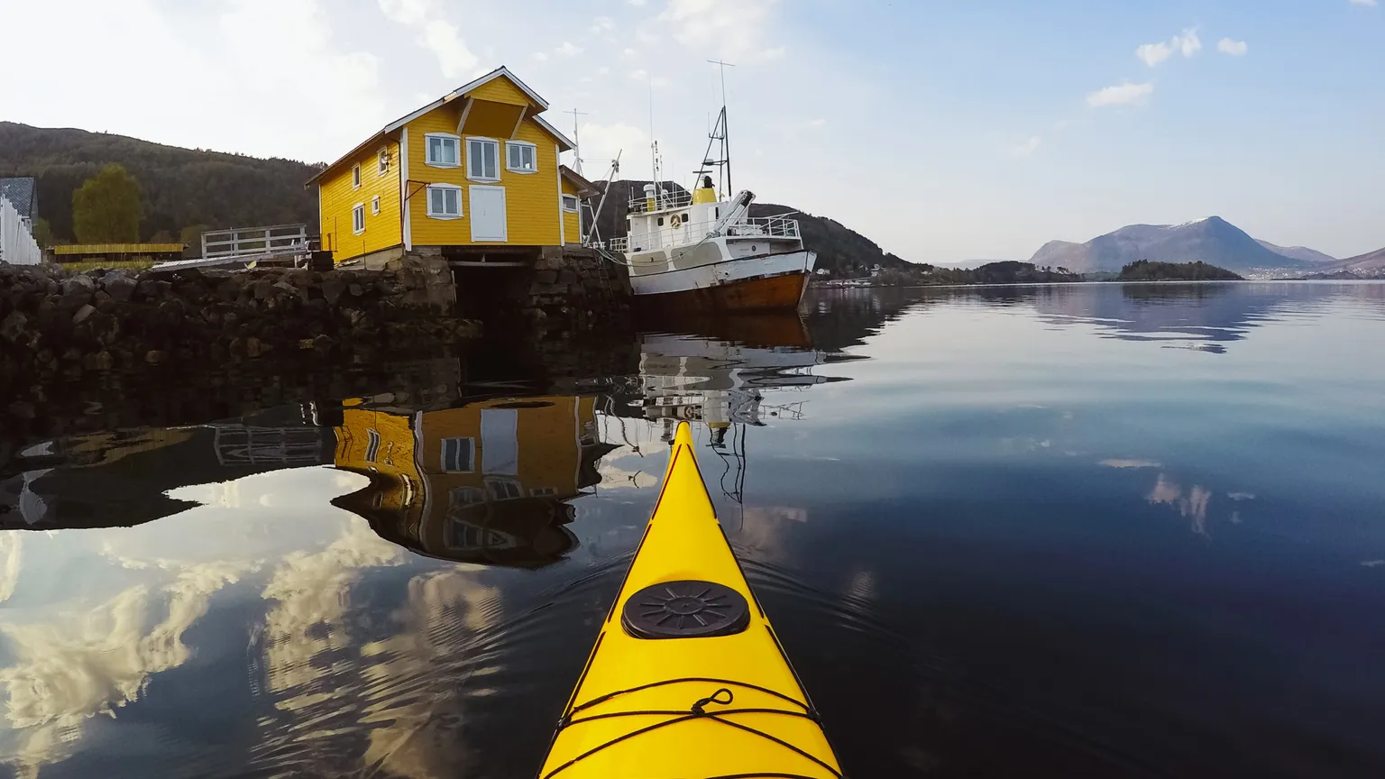 Kayak jaune sur l'eau, face à une maison jaune et un bateau sur la rive.