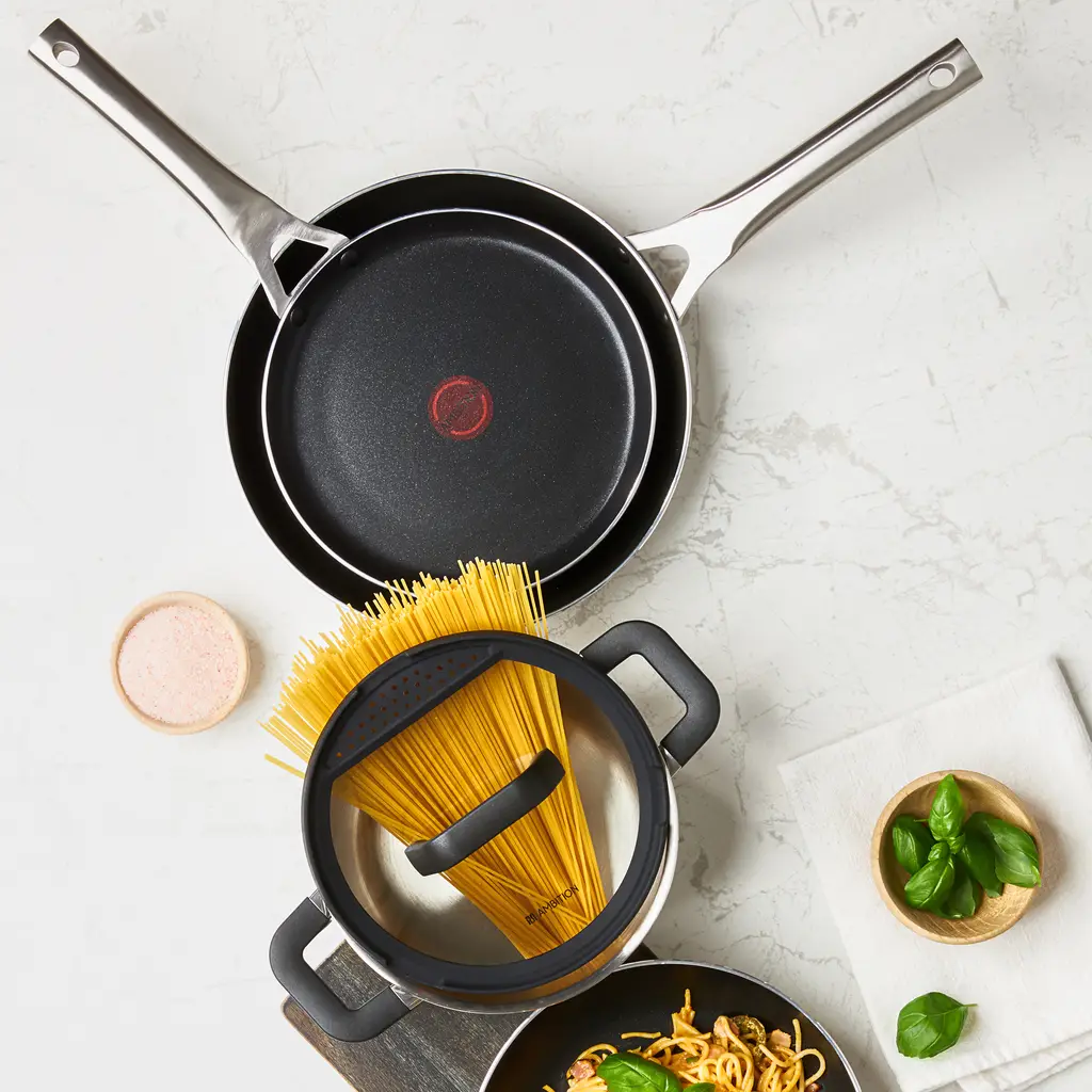 Scena gotowania z czarnymi patelniami, garnkiem ze spaghetti, bazylią i solą na marmurowej powierzchni.