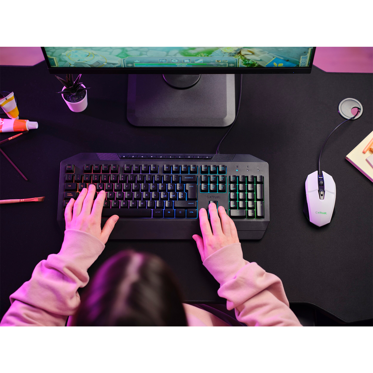 Mani su una tastiera con luce arcobaleno, schermo e mouse sulla scrivania.
