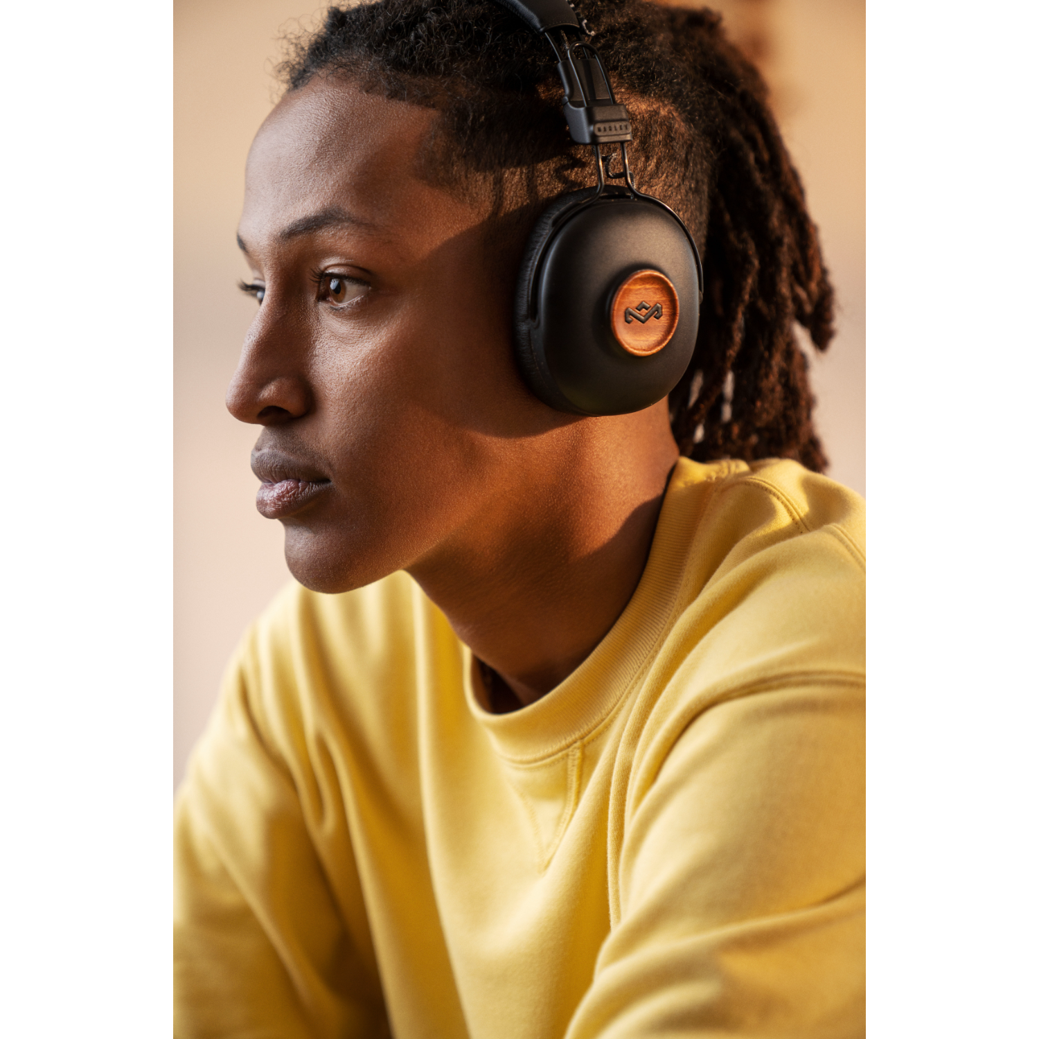 Jonge man met dreadlocks draagt zwarte koptelefoon met houten accenten, gele shirt, kijkt weg.