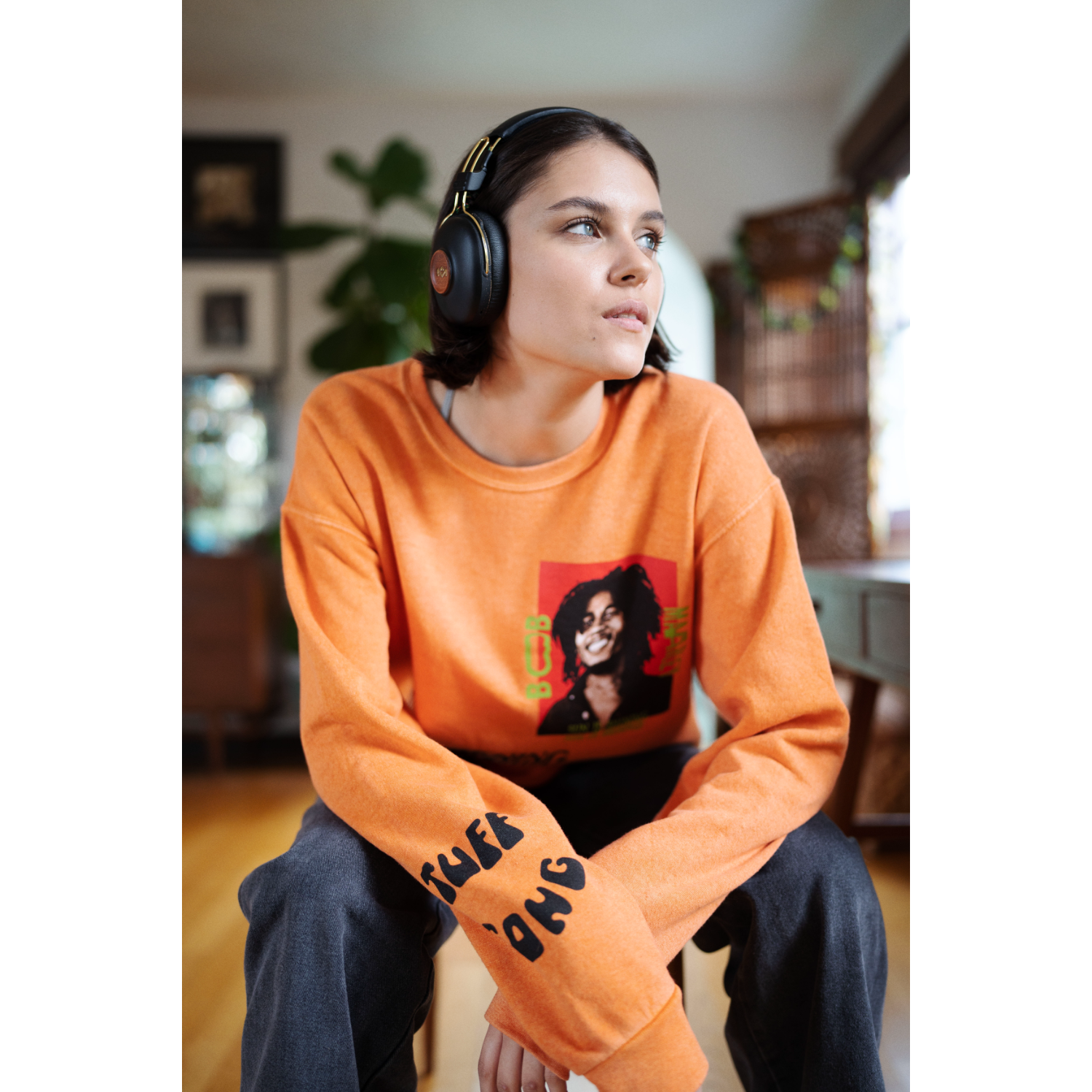 Woman wearing orange sweatshirt with Bob Marley image, black headphones, and dark pants, looking towards the right.