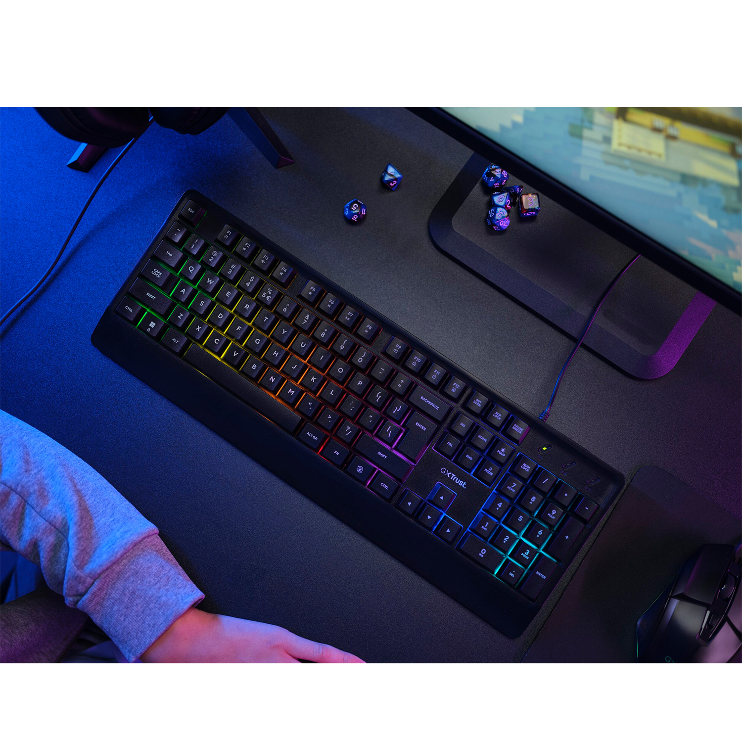 Tastiera gaming nera con tasti colorati su una scrivania con dadi e monitor.