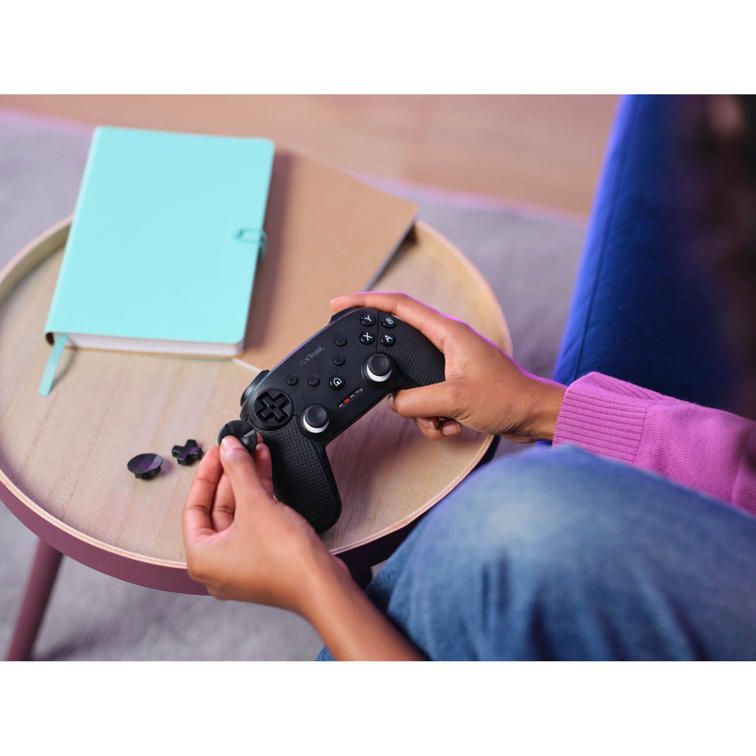 Le mani di una persona tengono un controller di gioco nero, con un quaderno blu e carta su un tavolino.