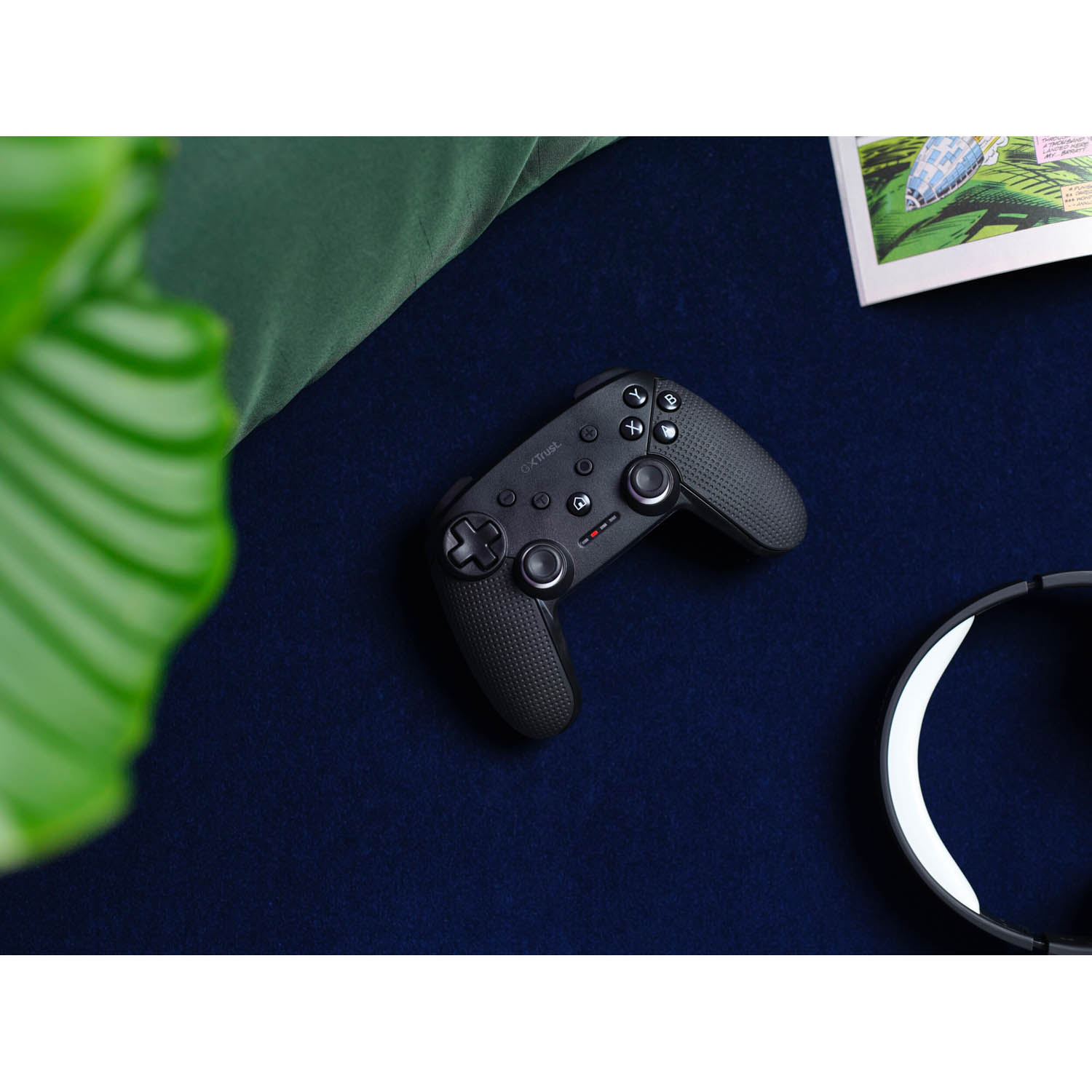 Controller di gioco nero su superficie blu. Pianta verde e fumetto visibili.