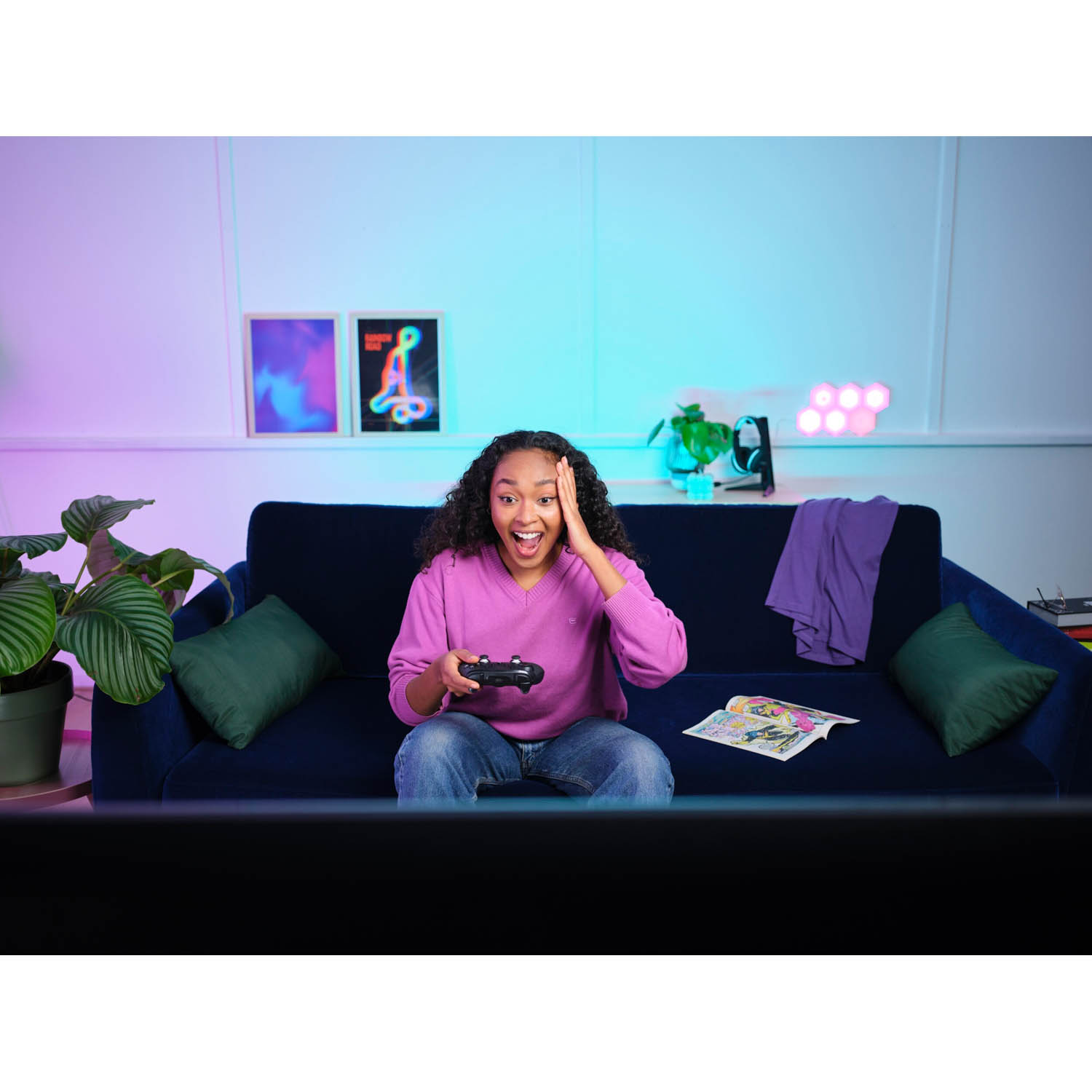 Una ragazza su un divano blu che gioca a un videogioco ed è sorpresa, illuminata da luci rosa e blu.