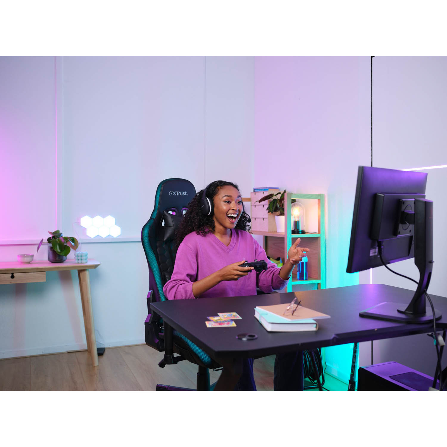 Donna che gioca. È eccitata, tiene un controller. Un monitor è visibile. Sedia e scrivania da gioco sono nella stanza.
