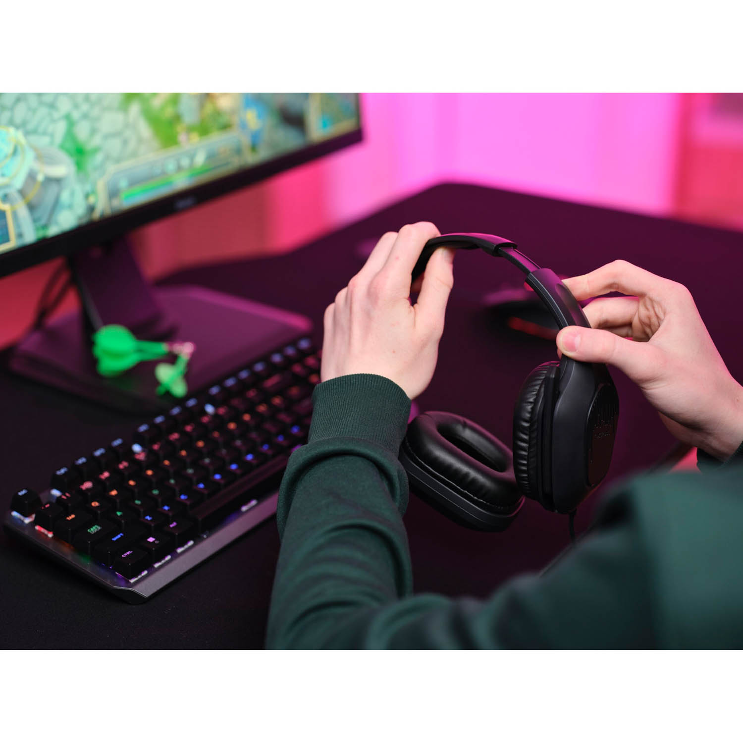 Persona che tiene le cuffie. Tastiera e monitor sullo sfondo. Figurina verde sulla scrivania.