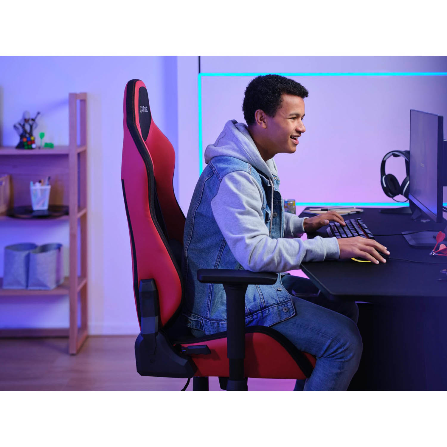 Uomo che gioca con sedia da gioco rossa, tastiera e monitor in una stanza con luci colorate.