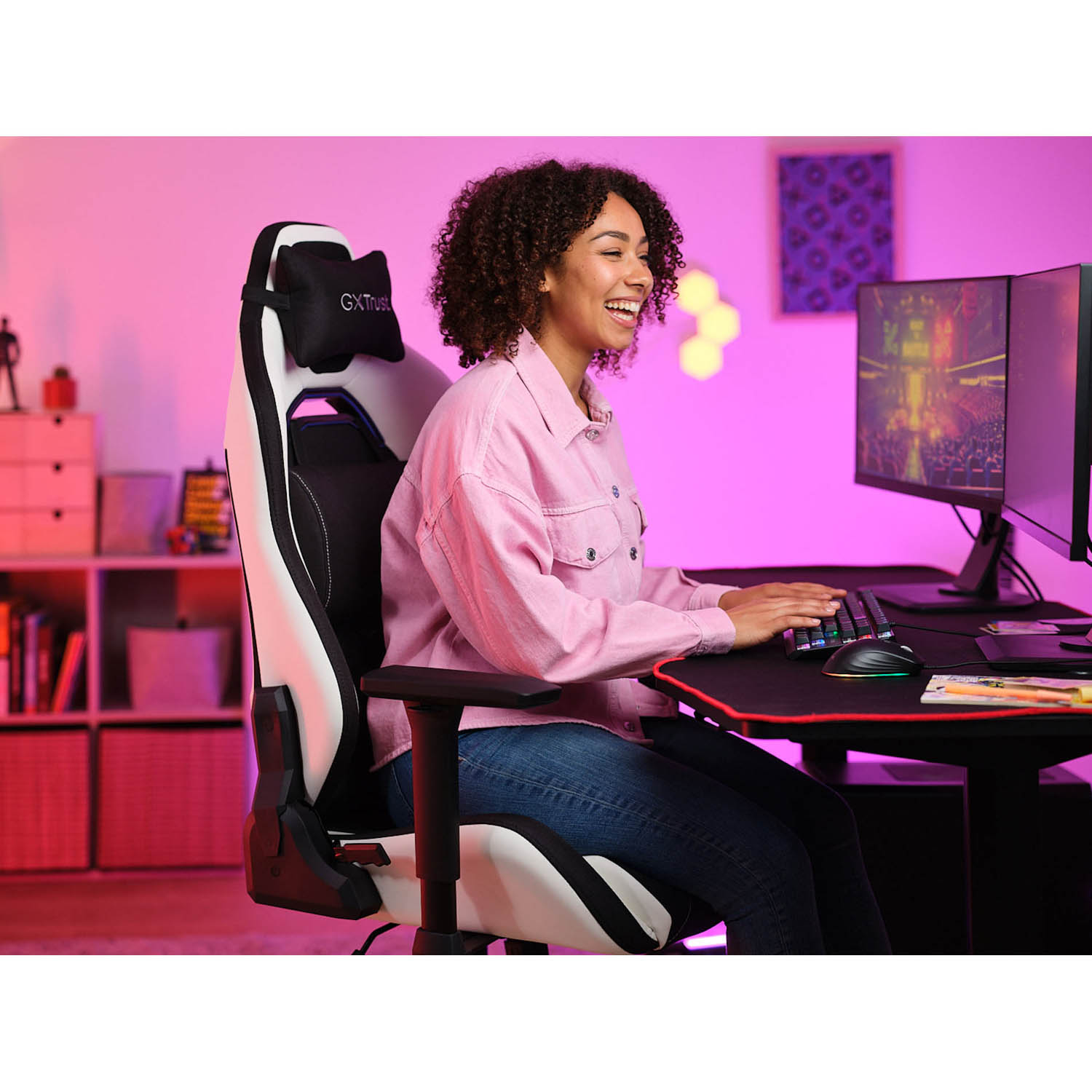 Donna in sedia da gaming con computer, tastiera e mouse. Illuminazione rosa e viola nella stanza.