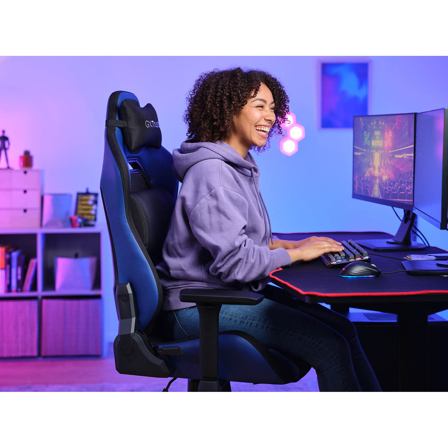 Una donna sorride mentre gioca. È su una sedia da gioco blu. La stanza è illuminata con luce blu e viola.