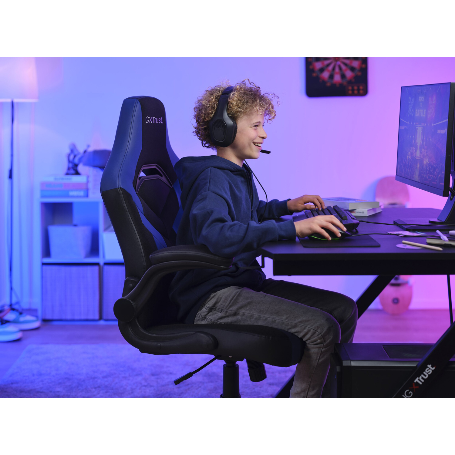 Un ragazzo con le cuffie gioca, seduto su una sedia da gaming nera e blu. Un monitor sullo sfondo.