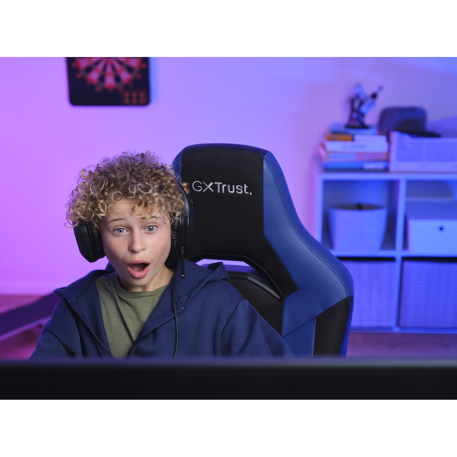 Ragazzo che gioca. Indossa le cuffie. Sedia e monitor da gioco. Espressione sorpresa. Luce blu.